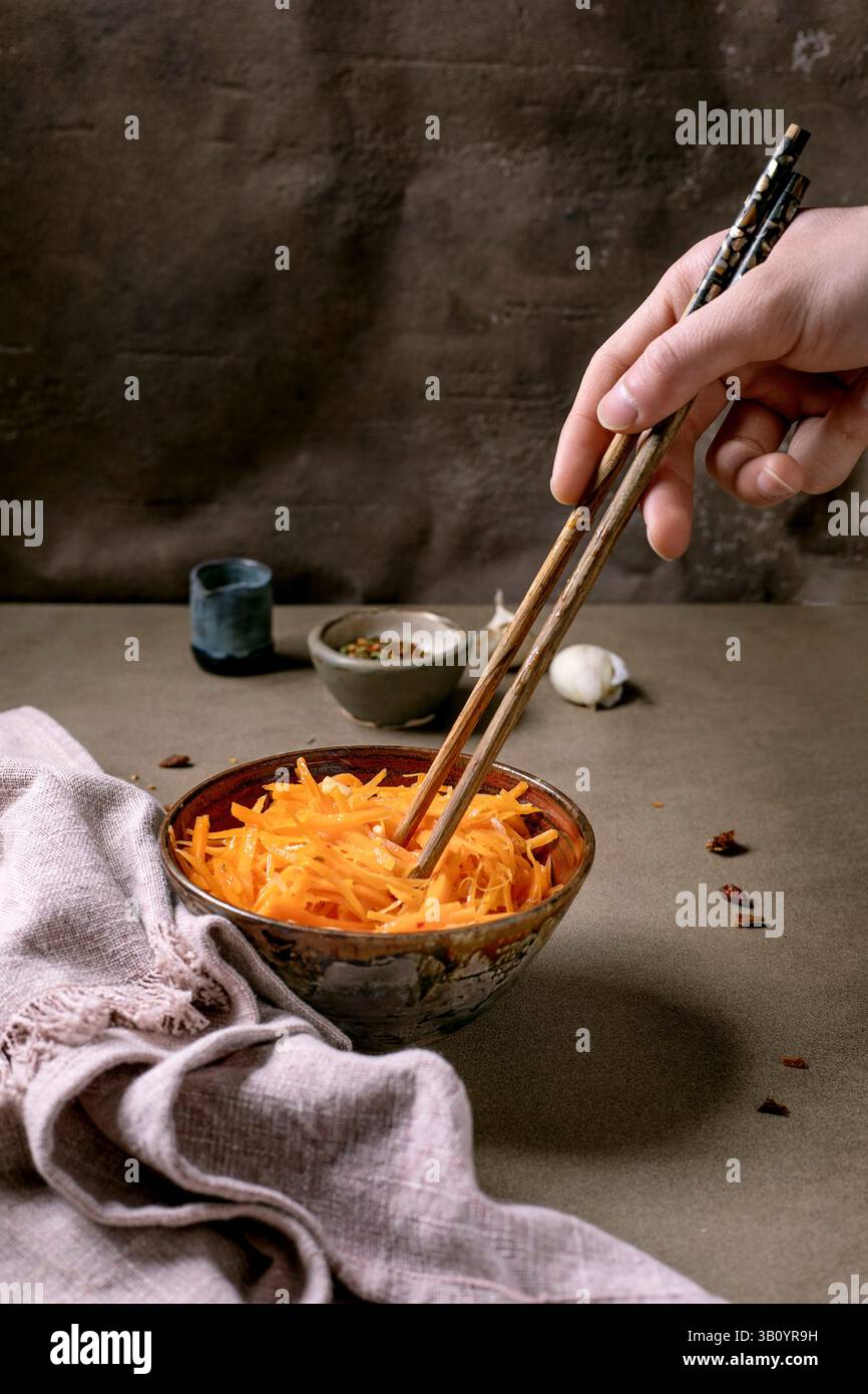 Koreanische und asiatische Küche. Die Hand des Mannes nimmt mit Stäbchen Gemüsesalat aus marinierten Karotten mit Knoblauch aus einer Keramikschale auf. Würzen in kleinen Größen Stockfoto