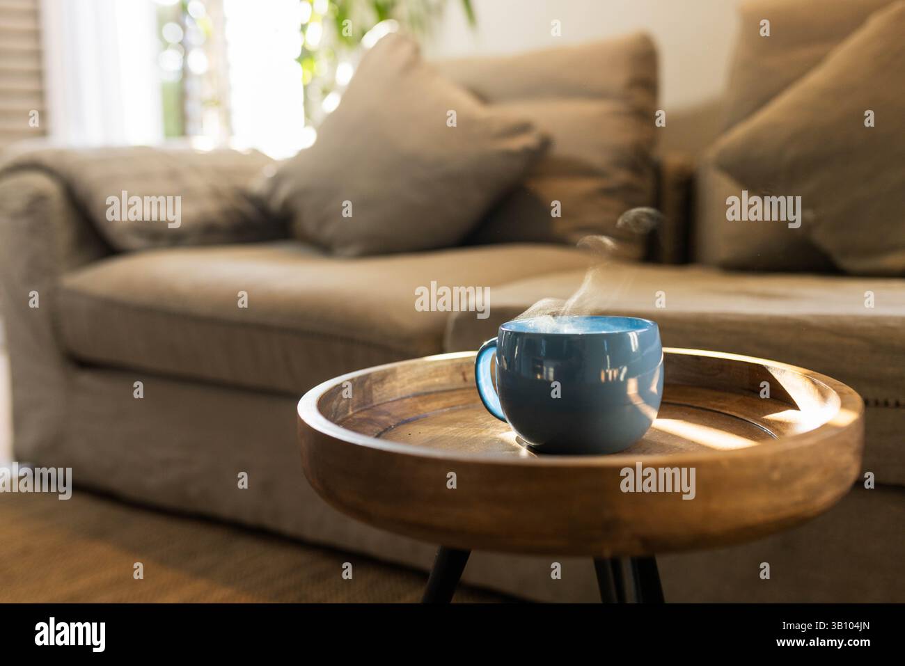 Dampfender blauer Becher auf rundem Holztisch mit beigefarbenem Sofa, Licht filtert durch weiße Jalousien Stockfoto
