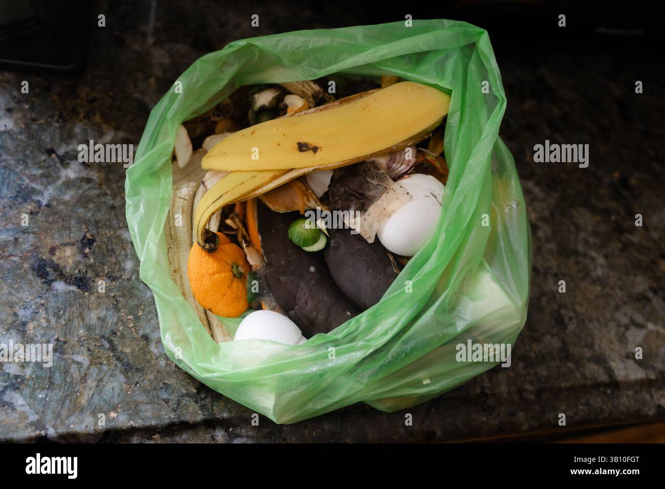 Verschiedene Obst- und Gemüsereste, die in einem grünen Beutel gesammelt werden, sitzen auf einer Küchenarbeitsfläche aus Granit. Stockfoto