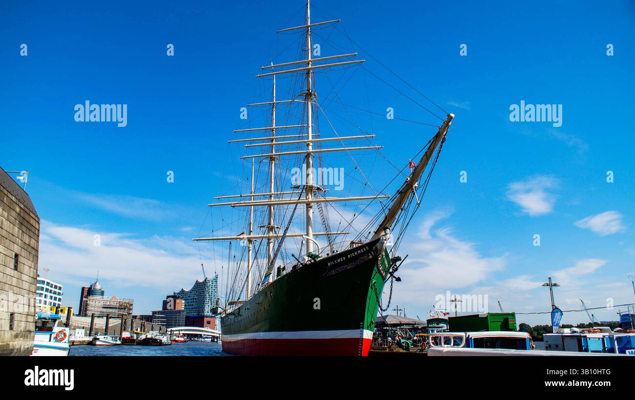 Ein wunderschön erhaltenes historisches Segelschiff, das in einem geschäftigen Hafen unter einem klaren blauen Himmel vor Anker liegt. Dieses elegante Seeschiff mit seinen hohen Masten Stockfoto