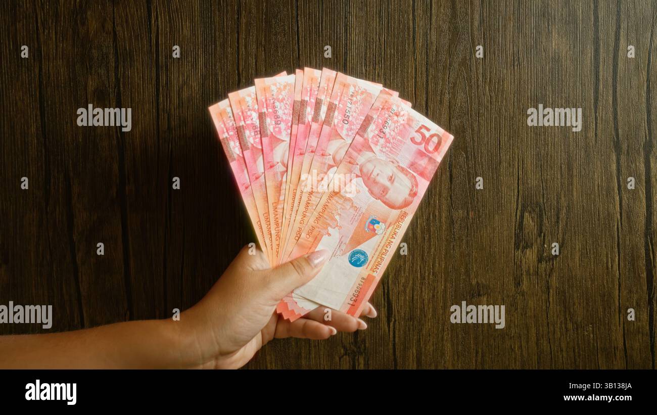 Frau, die mehrere philippinische Peso-Scheine vor einem rustikalen Holzhintergrund hält und Geldscheine mit sichtbaren Fingern in der Hand zeigt. Stockfoto