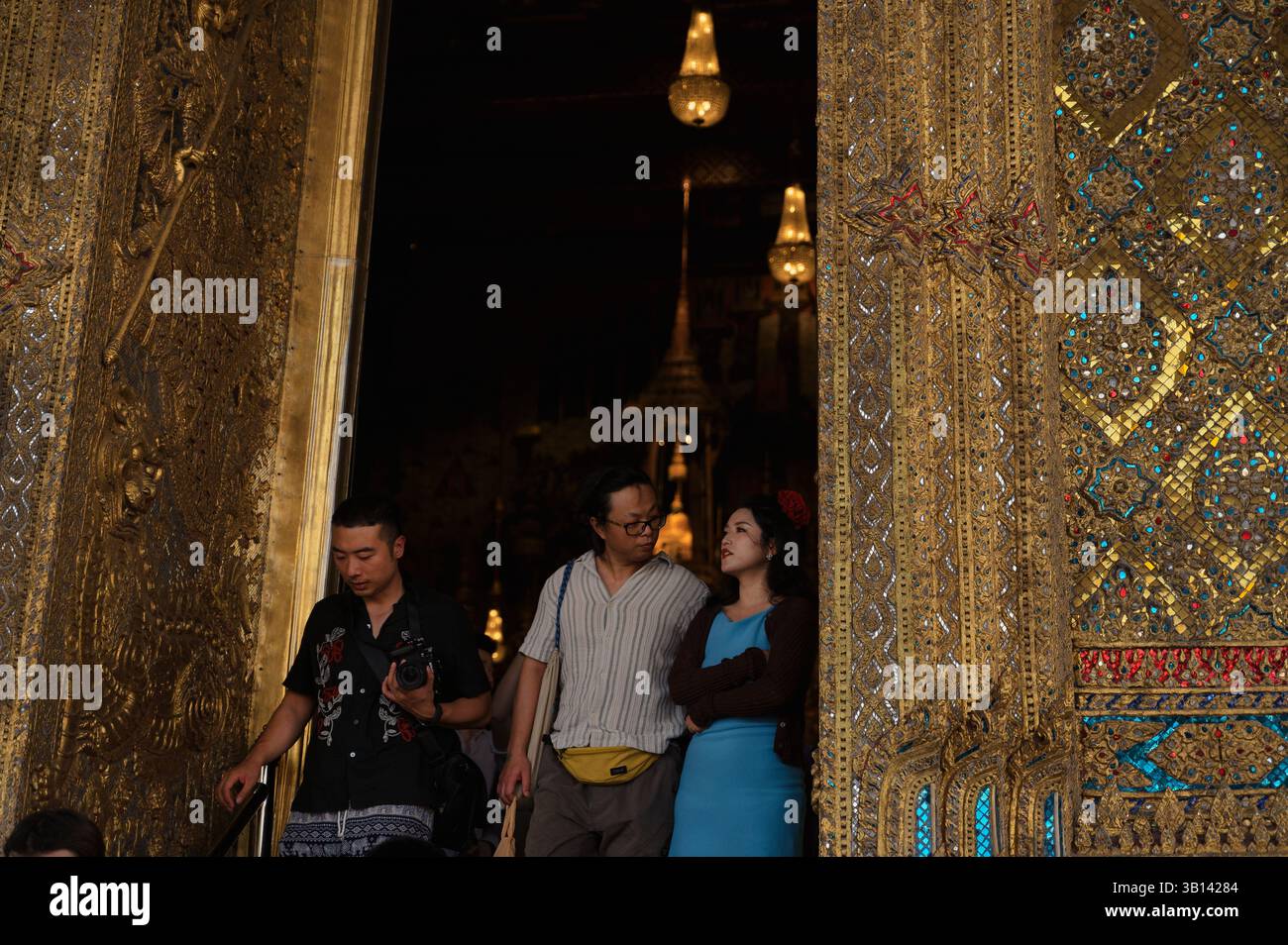 Touristen tauchen aus dem schwach beleuchteten Inneren des Wat Phra Kaew auf, eingerahmt von hohen goldenen Türen Stockfoto