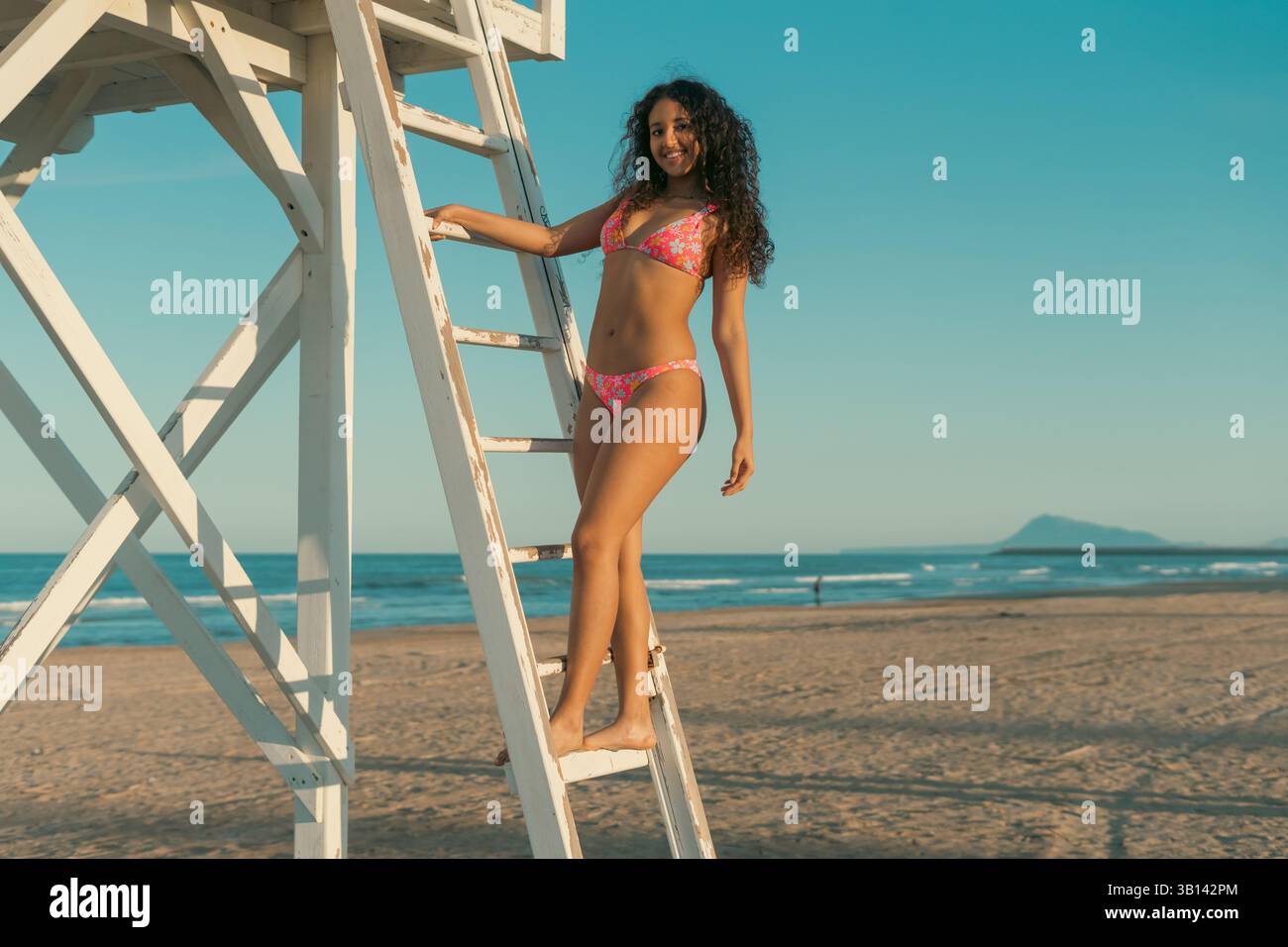 Lächelnde Frau in einem bunten Badeanzug, die auf einer Leiter am Meer steht. Der klare blaue Himmel und der ferne Berg bilden einen ruhigen Strand Stockfoto