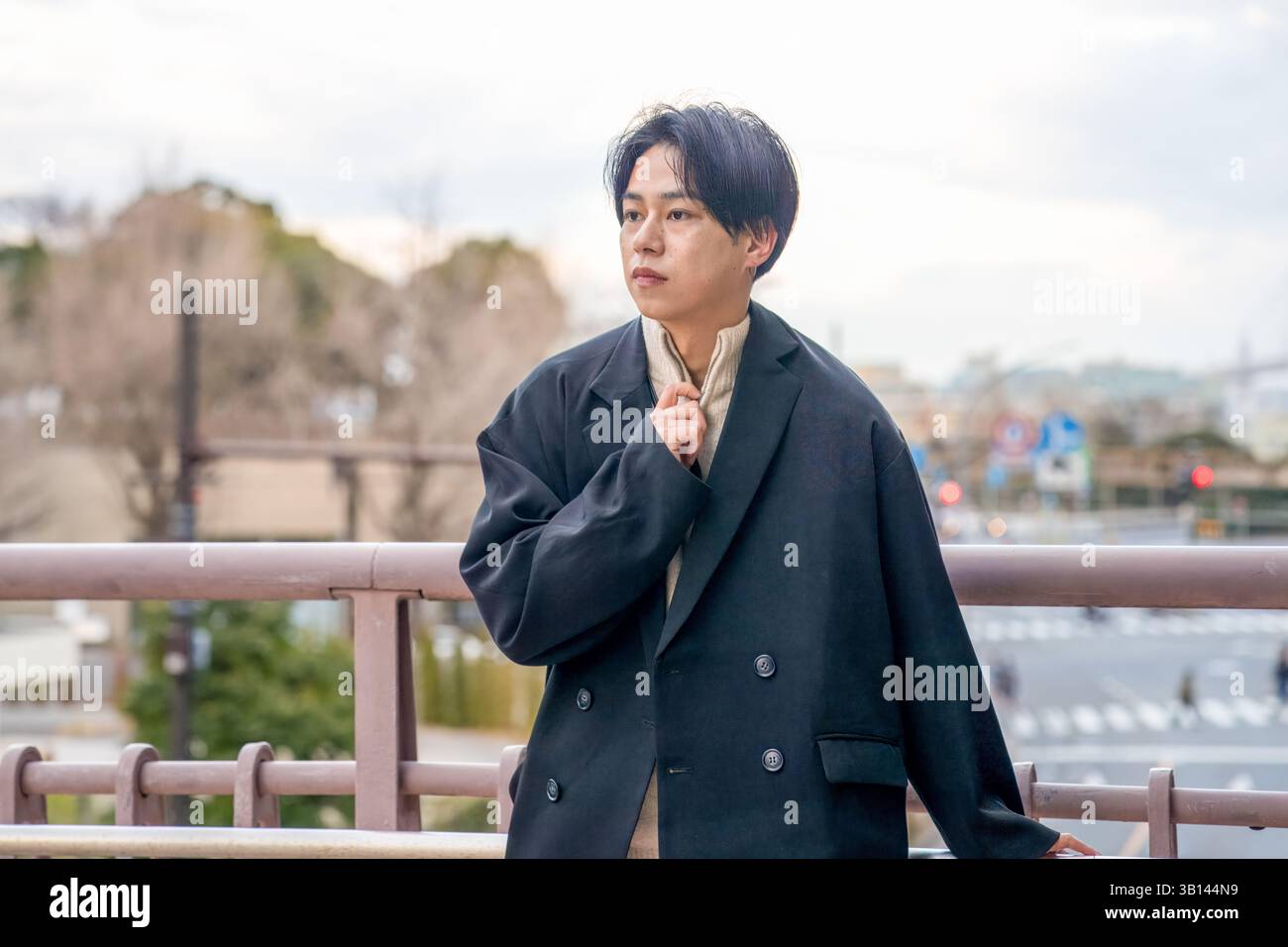 Ein Japaner in seinen Zwanzigern, der eine schwarze Jacke trägt, ist in Minato Mirai, Yokohama, Präfektur Kanagawa. Er sitzt auf einer Bank und blickt in die Ferne. Stockfoto