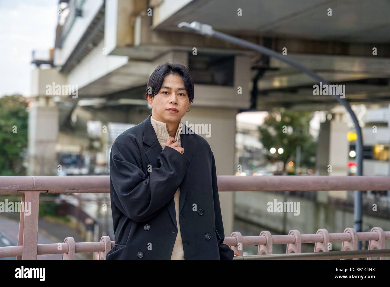 Ein Japaner in seinen Zwanzigern, der eine schwarze Jacke trägt, ist in Minato Mirai, Yokohama, Präfektur Kanagawa. Er sitzt auf einer Bank und blickt in die Ferne. Stockfoto