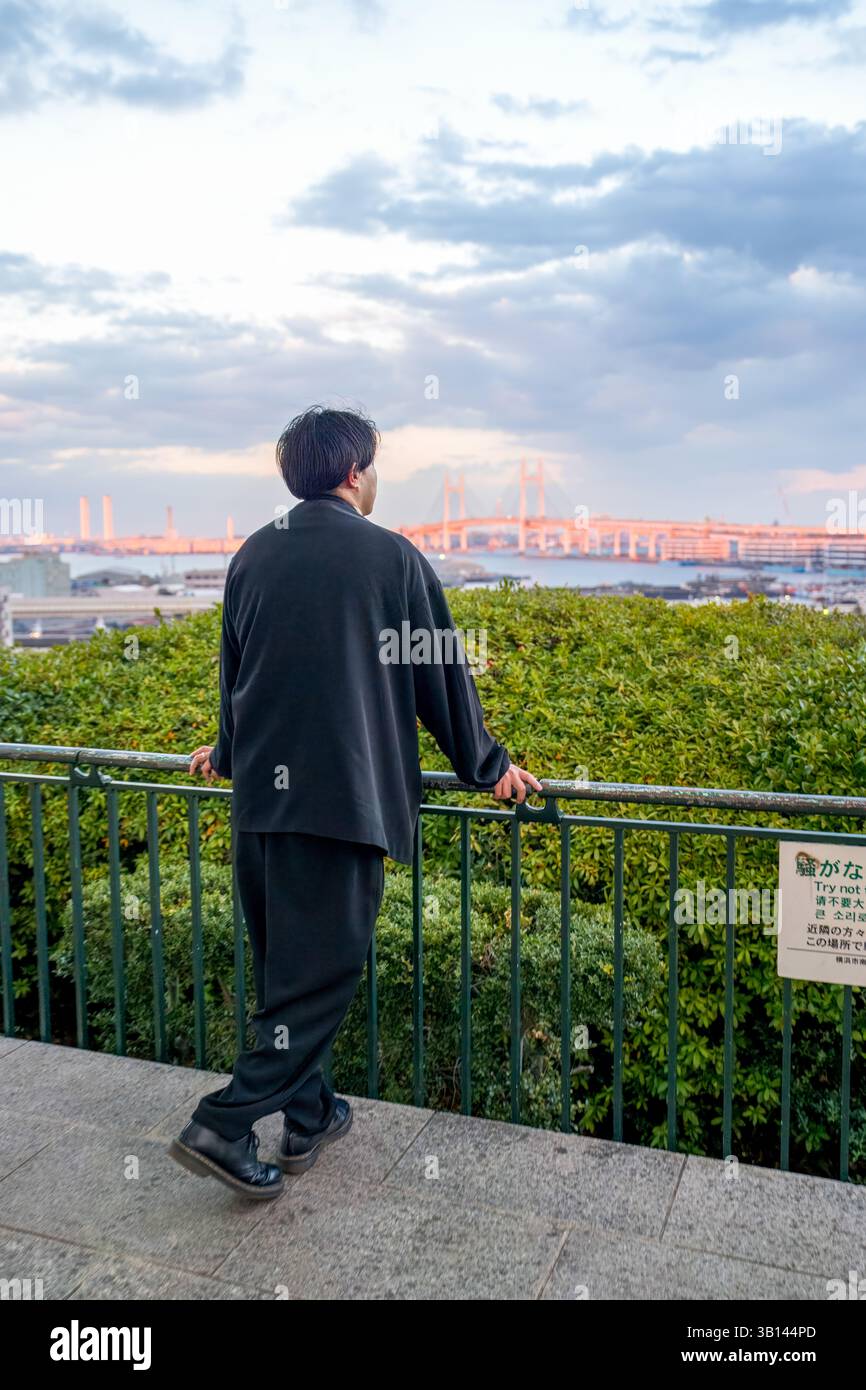 Ein Japaner in seinen Zwanzigern, der eine schwarze Jacke trägt, ist in Minato Mirai, Yokohama, Präfektur Kanagawa. Von hinten gesehen beobachtet er eine ferne Brücke Stockfoto