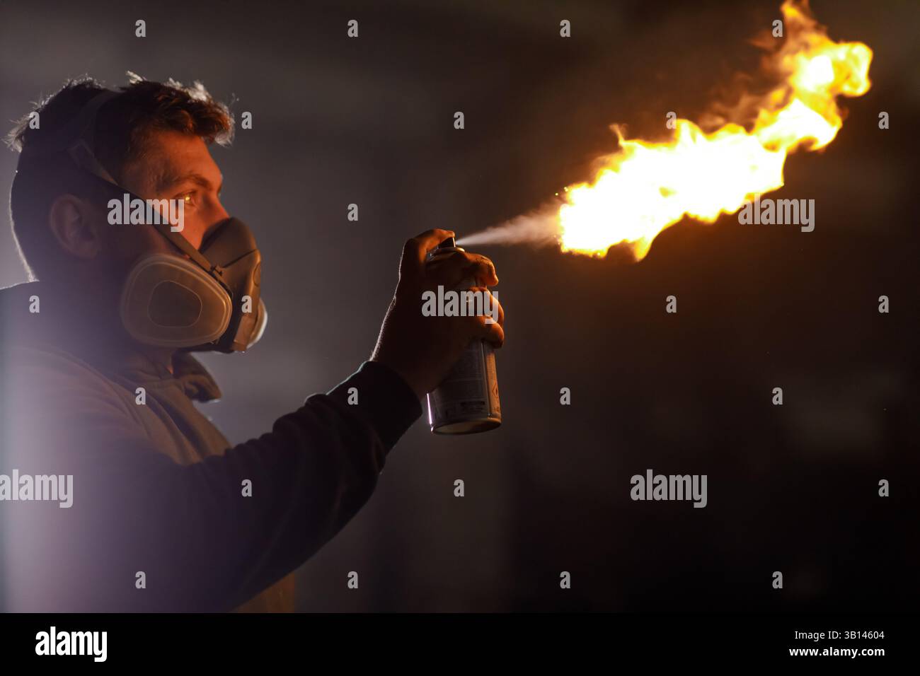 Eine Person in einer Schutzmaske zündet eine Sprühdose an und erzeugt in einer Werkstatt mit wenig Licht eine helle Flamme. Funken fliegen, während er sich sorgsam auf seine Aufgabe konzentriert Stockfoto
