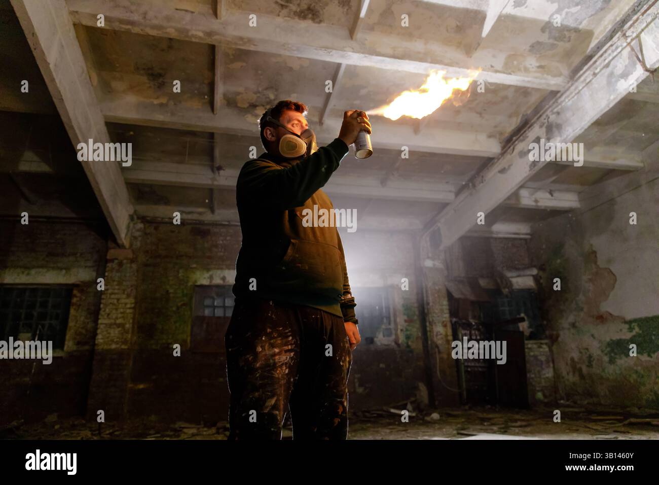 Eine Person in einem dunklen, verlassenen Industriebereich verwendet eine Sprühdose, um eine Flamme zu erzeugen. Die dunkle Umgebung hebt das Licht des Feuers hervor und zeigt den Schein Stockfoto