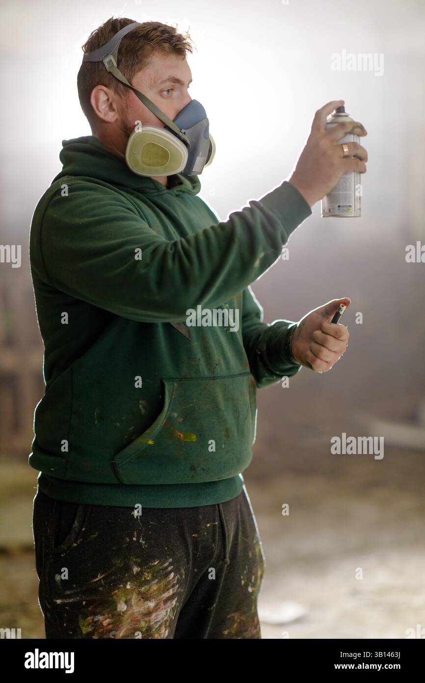 Ein Künstler verwendet Sprühfarbe, um ein buntes Stück in einer Werkstatt zu kreieren, die mit Kunstgegenständen gefüllt ist. Er trägt einen grünen Hoodie und eine grüne Maske und konzentriert sich auf seinen cr Stockfoto