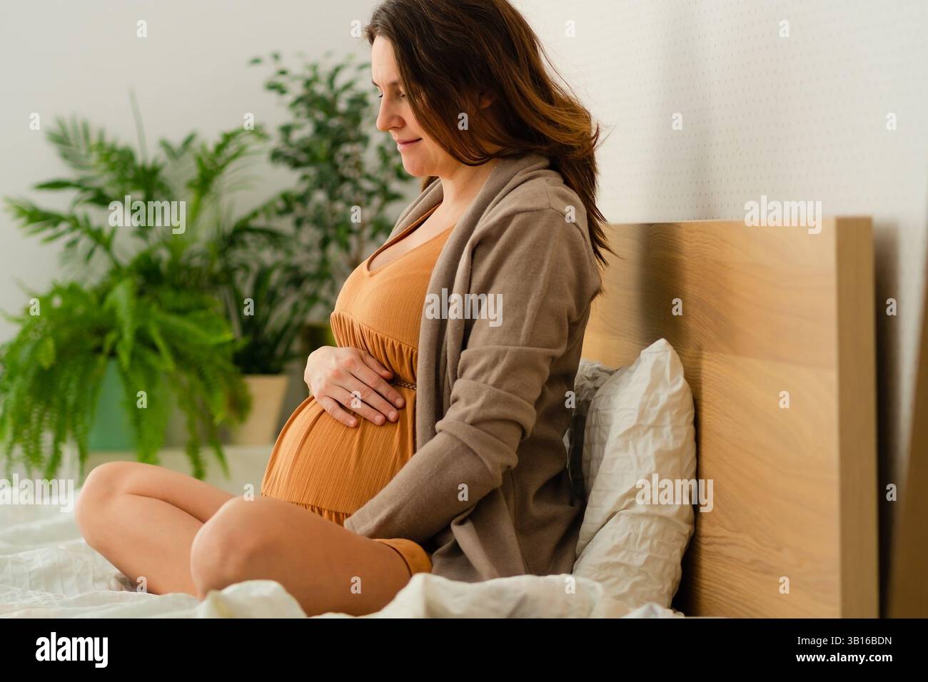 Eine heitere schwangere Frau sitzt in Lotusposition auf einem gemütlichen Bett und streichelt ihren Bauch sanft. Die warme Atmosphäre spiegelt die freudige Erwartung der Mutterschaft wider, und sanfte Beleuchtung verstärkt den emotionalen Moment. Stockfoto