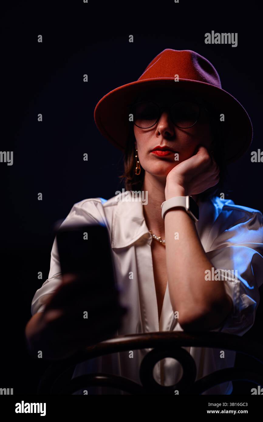 Die modische Frau mit rotem Hut und Satinbluse legt ihren Kopf auf ihre Hand, während sie bei schlechten Lichtverhältnissen auf ihr Handy blickt. Stockfoto