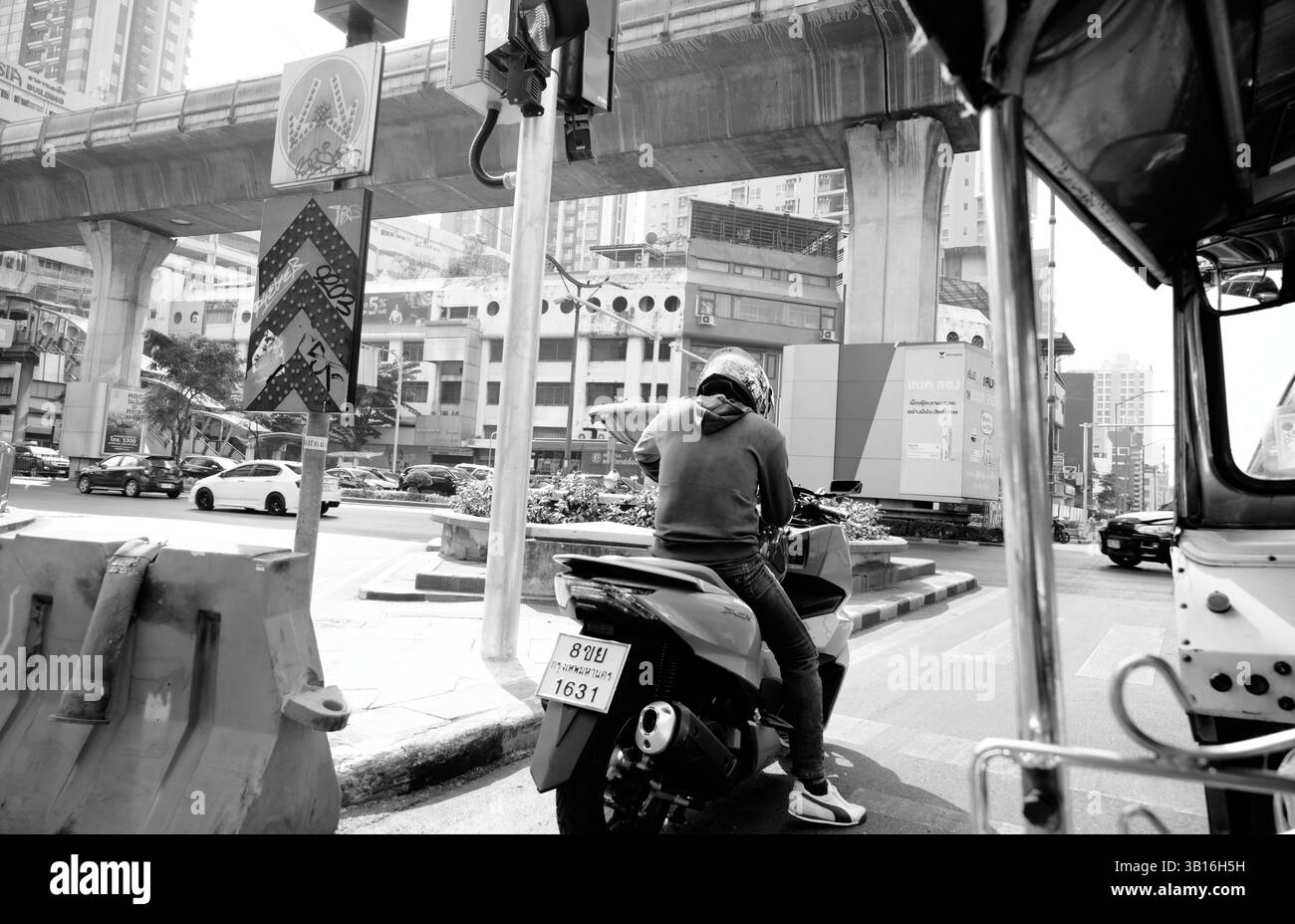 Der Motorradfahrer wartet an einer geschäftigen Kreuzung, eingerahmt von hohen Überführungen und dem Rücksitz eines Tuk-Tuk Stockfoto