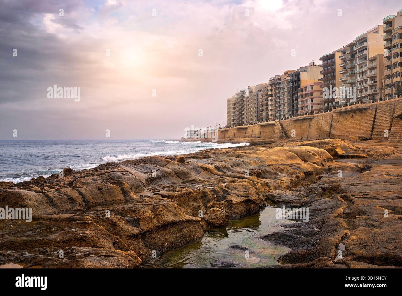 Felsige Küste mit Apartmenthäusern entlang einer hohen Promenade. Weiches Licht bei Sonnenuntergang hebt die Texturen der Felsen und des Meeres hervor. Friedliche Küstenatmosphäre Stockfoto