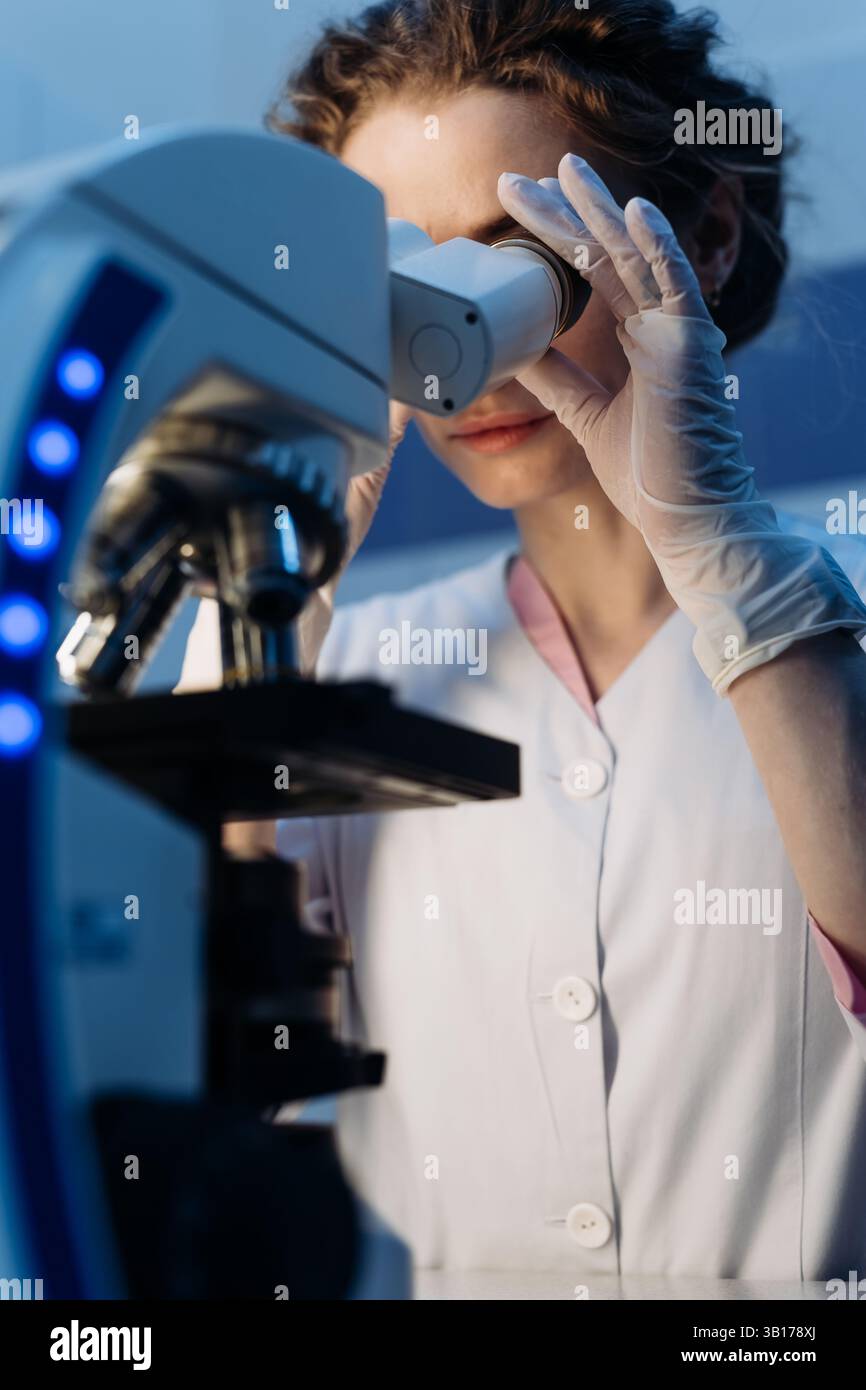 Wissenschaftlerin in steriler Kleidung, die am Tisch sitzt und Forschungsuntersuchungen in einem medizinischen Labor mit einem Mikroskop durchführt. Mikrobiologie Stockfoto