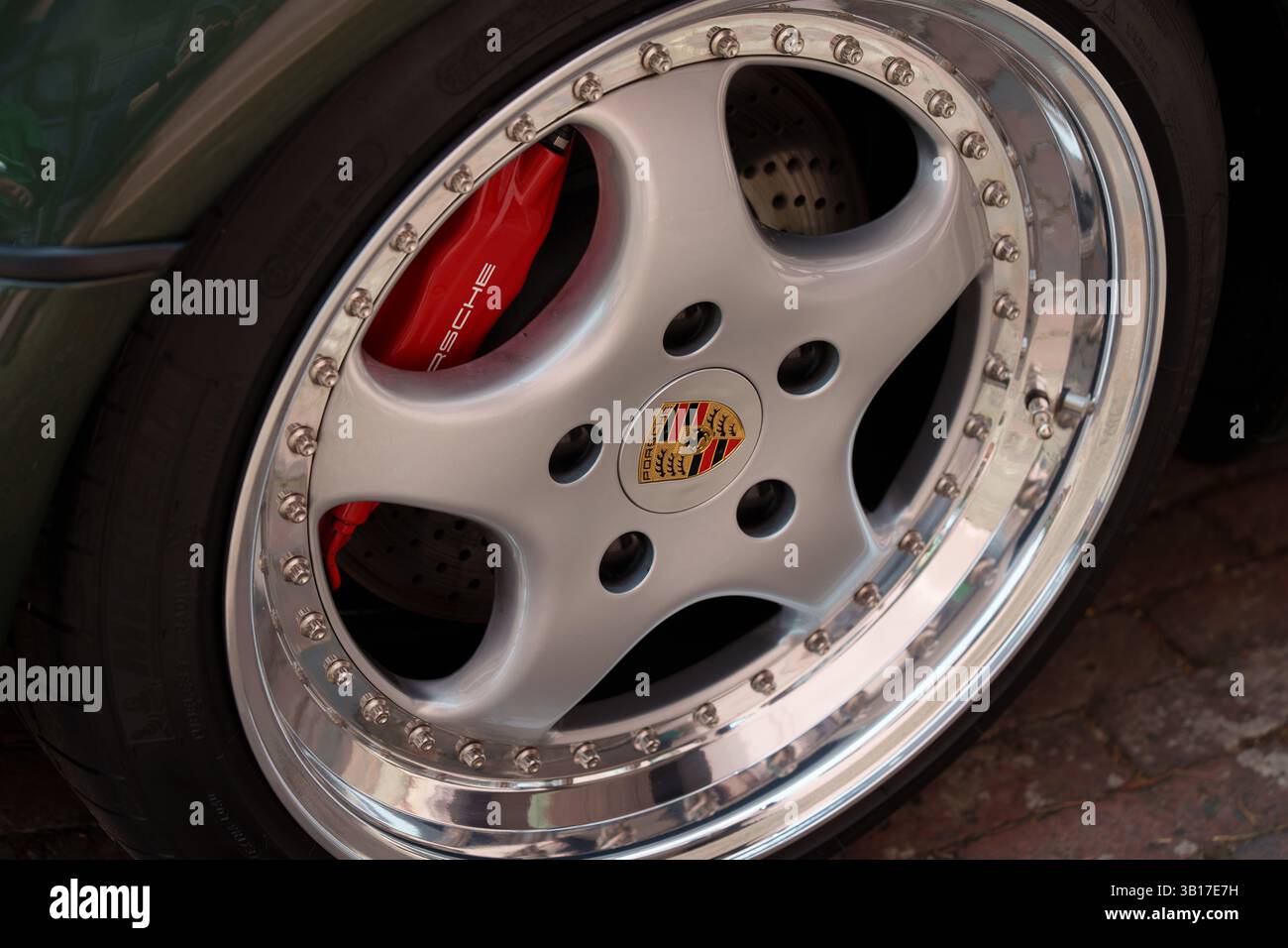 Nahaufnahme einer silbernen Porsche-Leichtmetallfelge mit rotem Bremssattel und polierter Chromfelge, die auf einem Ziegelpflaster geparkt ist. Stockfoto