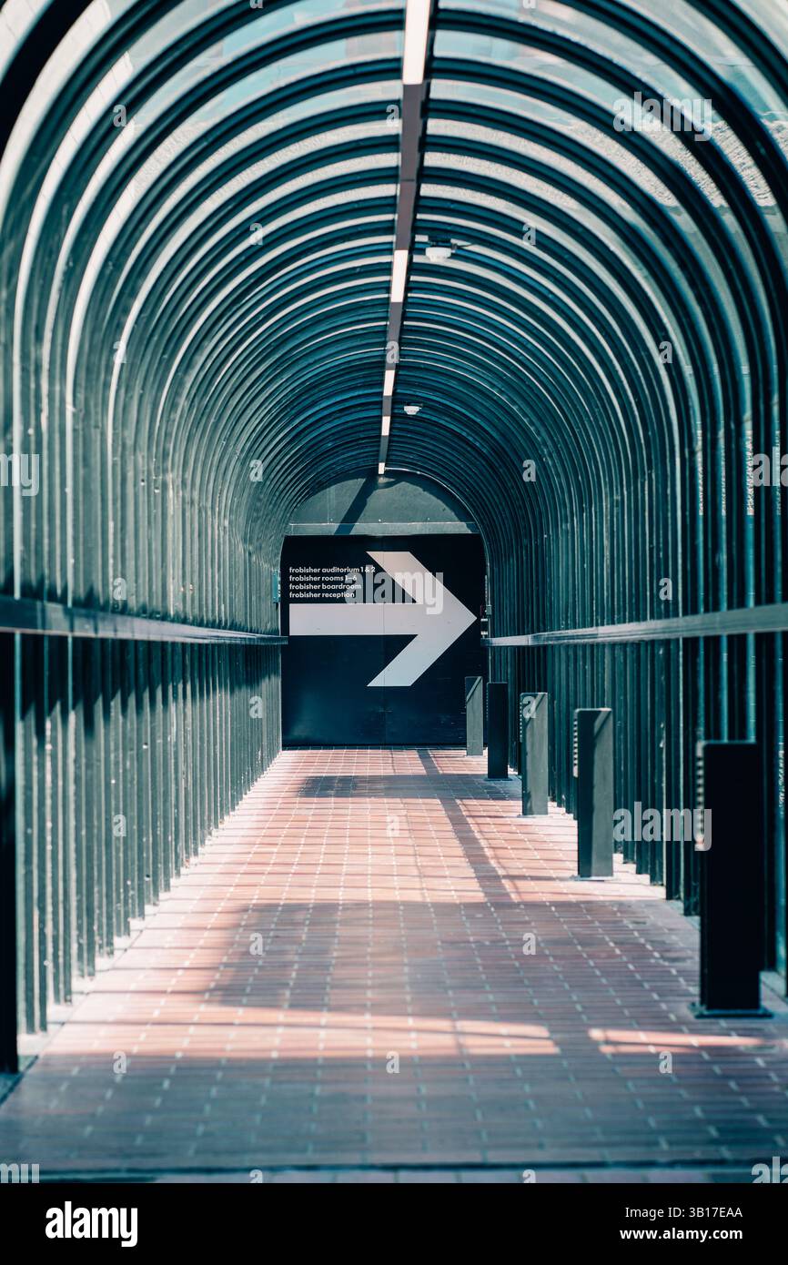 Ein überdachter Gang mit Ziegelboden führt zu einer dunklen Tür mit einem weißen Pfeil, der auf den Weg zeigt, was ein Gefühl der Richtung und des Geheimnisses schafft. Stockfoto