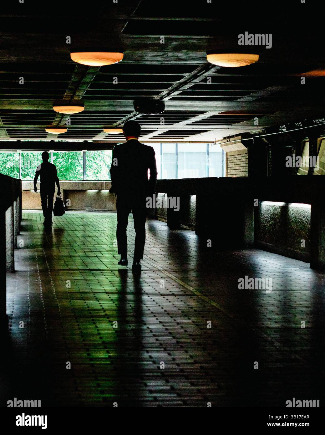 Zwei Männer gehen durch eine schwach beleuchtete, moderne Unterführung, deren Silhouetten sich von dem hellgrünen Laub abheben, das durch ein Fenster sichtbar ist. Stockfoto