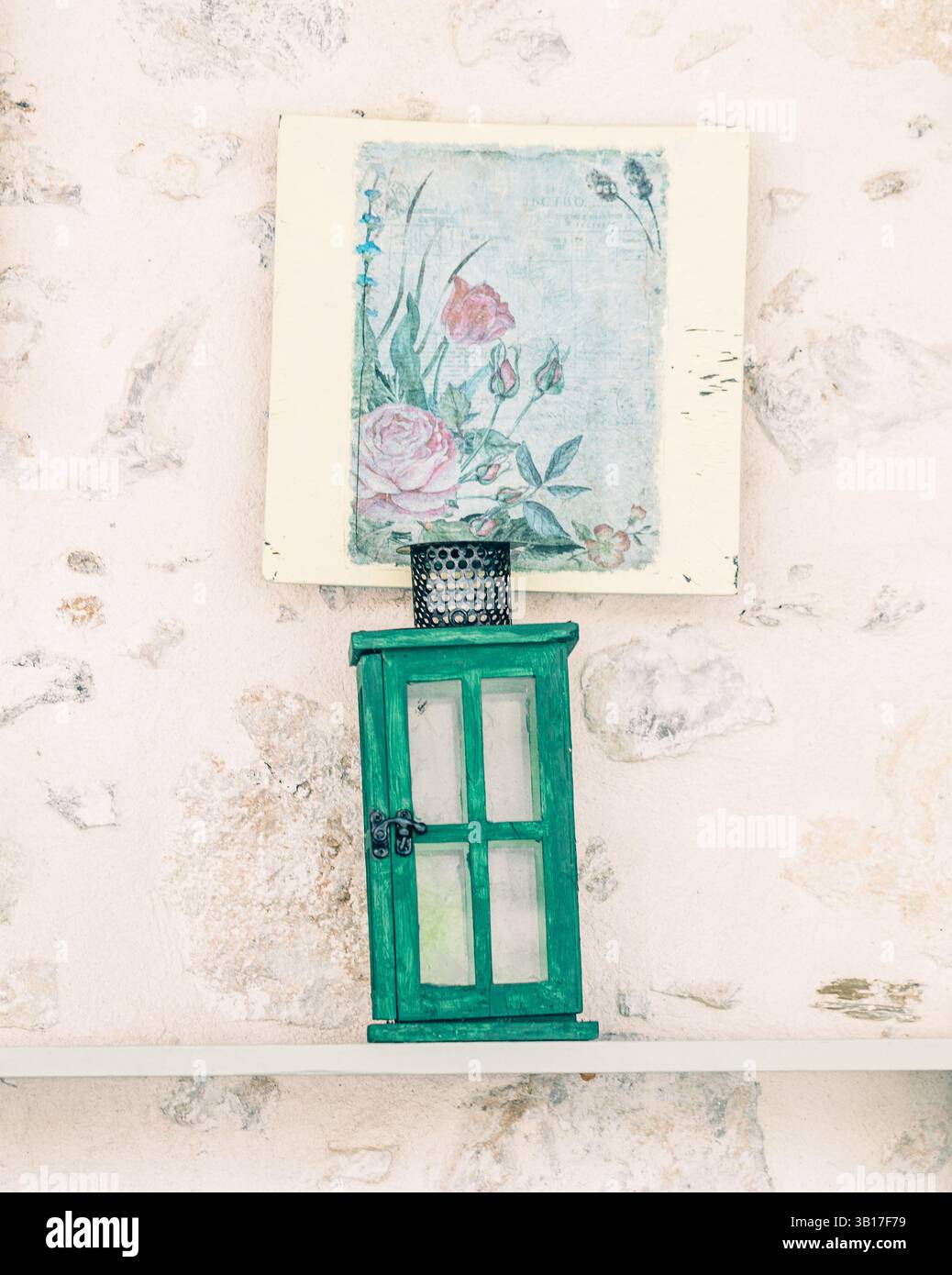 Eine grüne hölzerne Laterne sitzt auf einem Regal unter einem gerahmten Blumenmuster vor einer strukturierten Steinwand und schafft ein rustikales, Vintage-Stillleben in Griechenland. Stockfoto