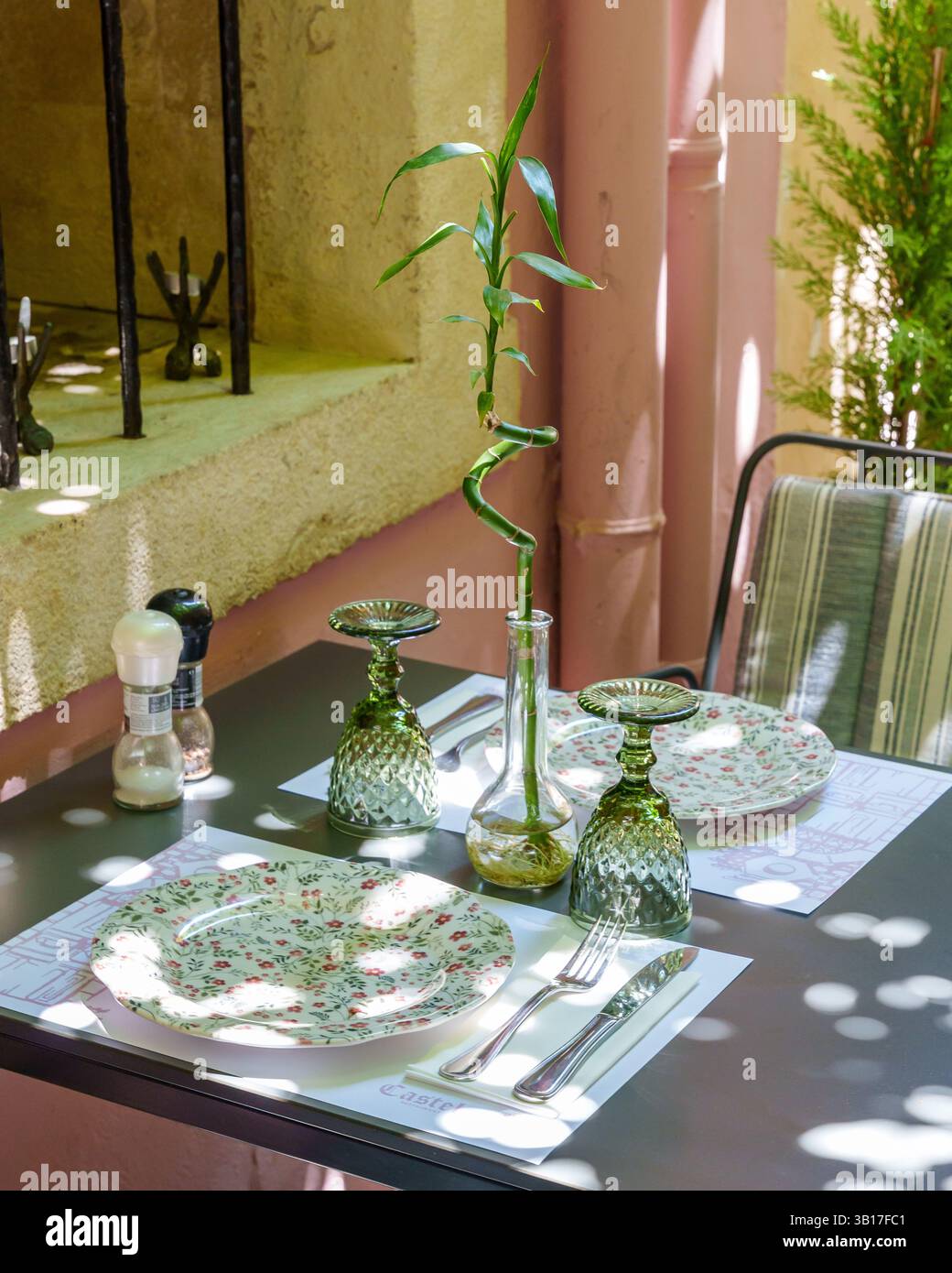 Ein bezauberndes Restaurant im Freien, das in strahlendes Sonnenlicht getaucht ist, mit Blumenteller, grünen Gläsern und einem Herzstück aus Bambus in Griechenland. Stockfoto