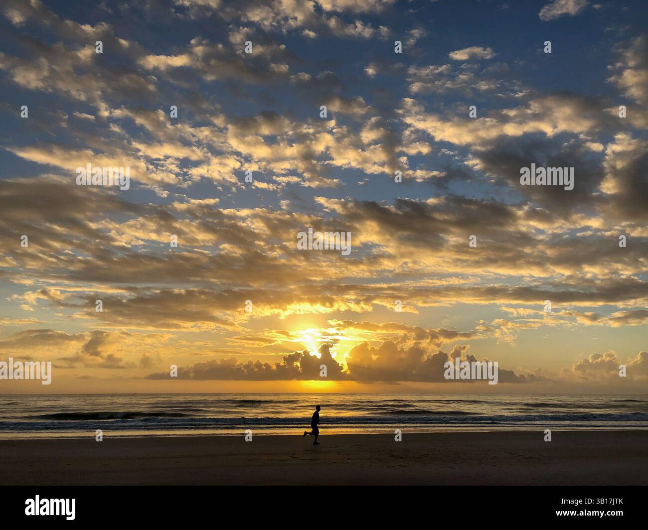 Silhouette eines Mannes, der bei Sonnenaufgang am Strand läuft, mit dramatischen Wolken über dem Meer. Amelia Island, Florida, USA. - Smartphone-aufgenommenes Stockfoto