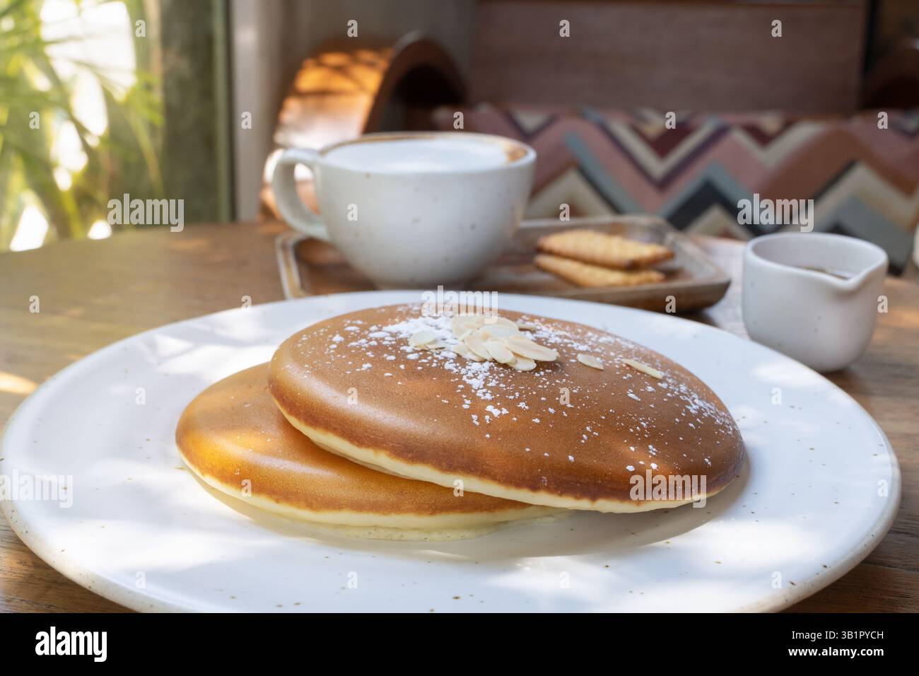 Ein gemütlicher Morgen in einem sonnigen Café. Ein Stapel flauschiger hausgemachter Pfannkuchen sitzt auf einem weißen Teller, gekrönt mit einem Spritzer süßen Sirup. Servierte Alongs Stockfoto