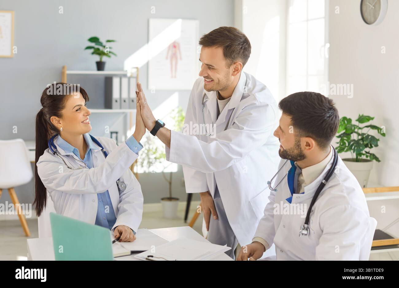 Drei lächelnde Ärzte feiern erfolgreiche Arbeit und geben hohe fünf an Kollegen in der medizinischen Praxis. Stockfoto