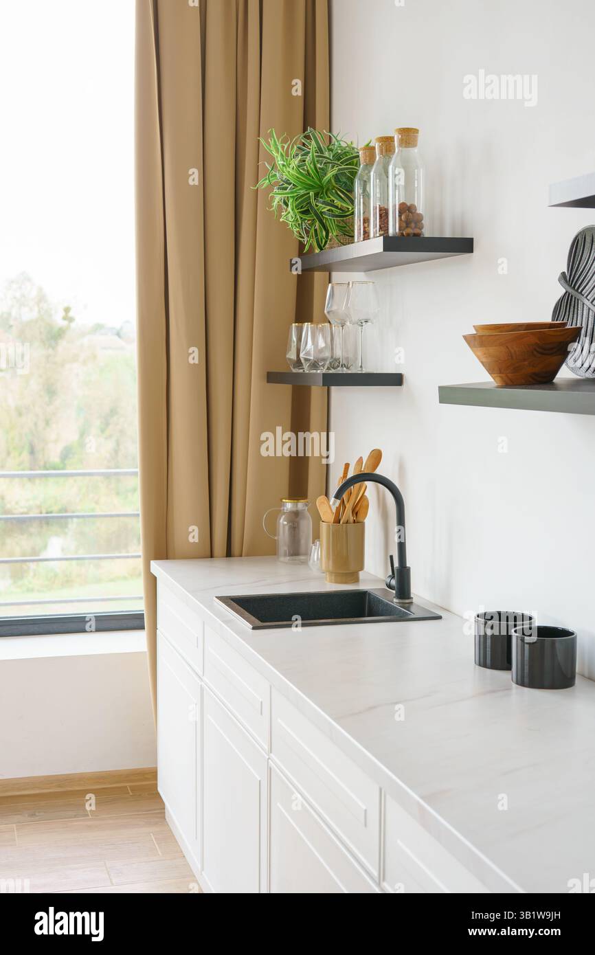 Weiße Küche mit Arbeitsplatte und schwarzem Wasserhahn auf weißem Wandhintergrund mit Holzregalen. Regale mit Utensilien, Küchenschrank und Einrichtung. Design in Stockfoto