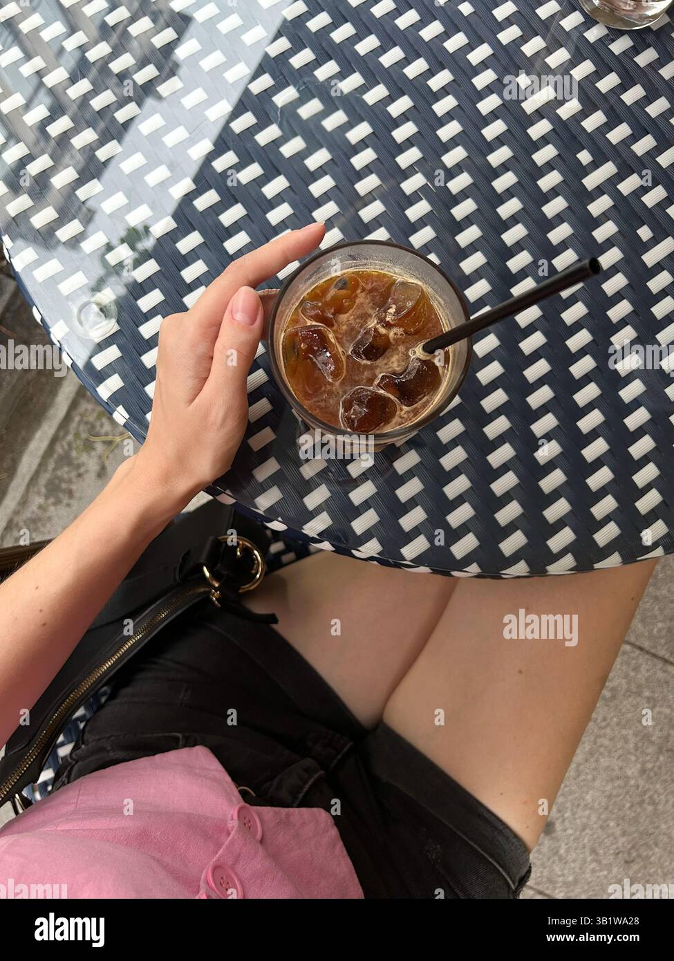 Das Foto zeigt eine Person, die ein Glas Eiskaffee hält, gefüllt mit Eiswürfeln und einem Strohhalm, an einem gewebten, blau-weiß gemusterten Tisch sitzt. Die Pers Stockfoto