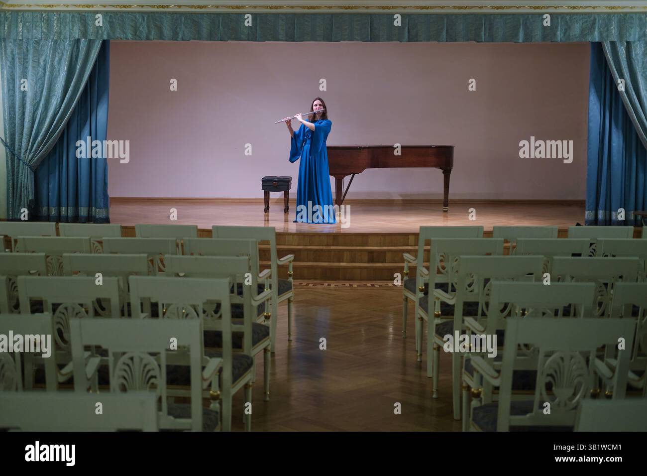 Flötist in blauem Kleid tritt auf einer leeren Konzertbühne mit einem Klavier im Hintergrund auf. Die ruhige Umgebung unterstreicht die emotionale Tiefe des so Stockfoto