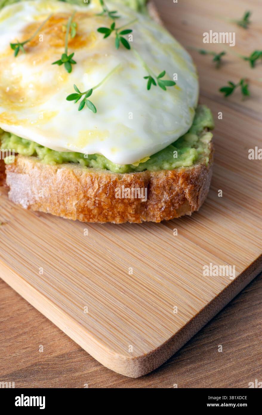 Ein Stück geröstetes Brot mit Avocadopüree, Spiegelei und frischen grünen Kressen auf einem Holzbrett Stockfoto