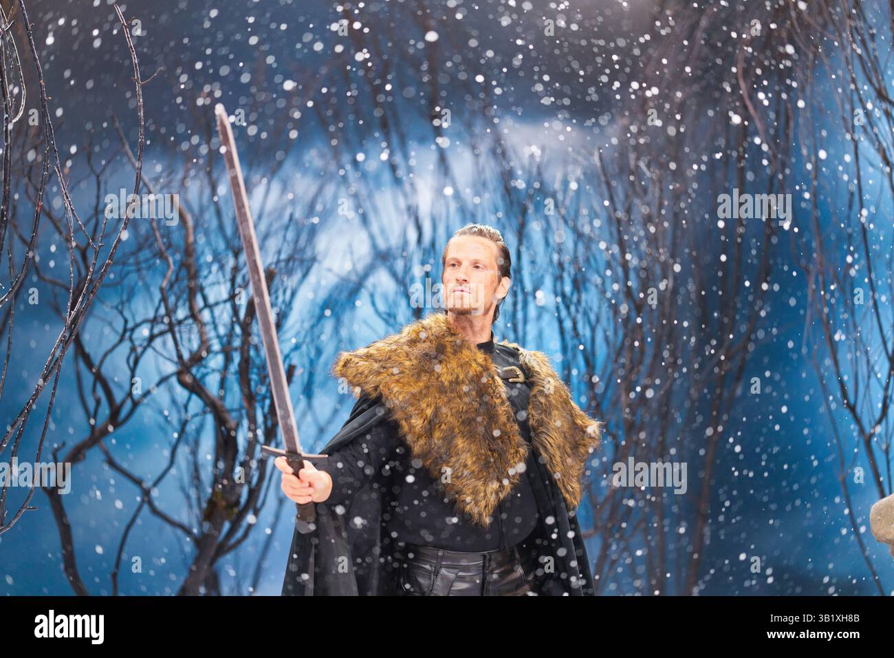 Ein hellhäutiger Mann mit kurzen braunen Haaren, trägt einen schwarzen Mantel und einen Pelzkragen, hält ein langes Schwert. Er steht in einem verschneiten, winterlichen Wald Agave Stockfoto