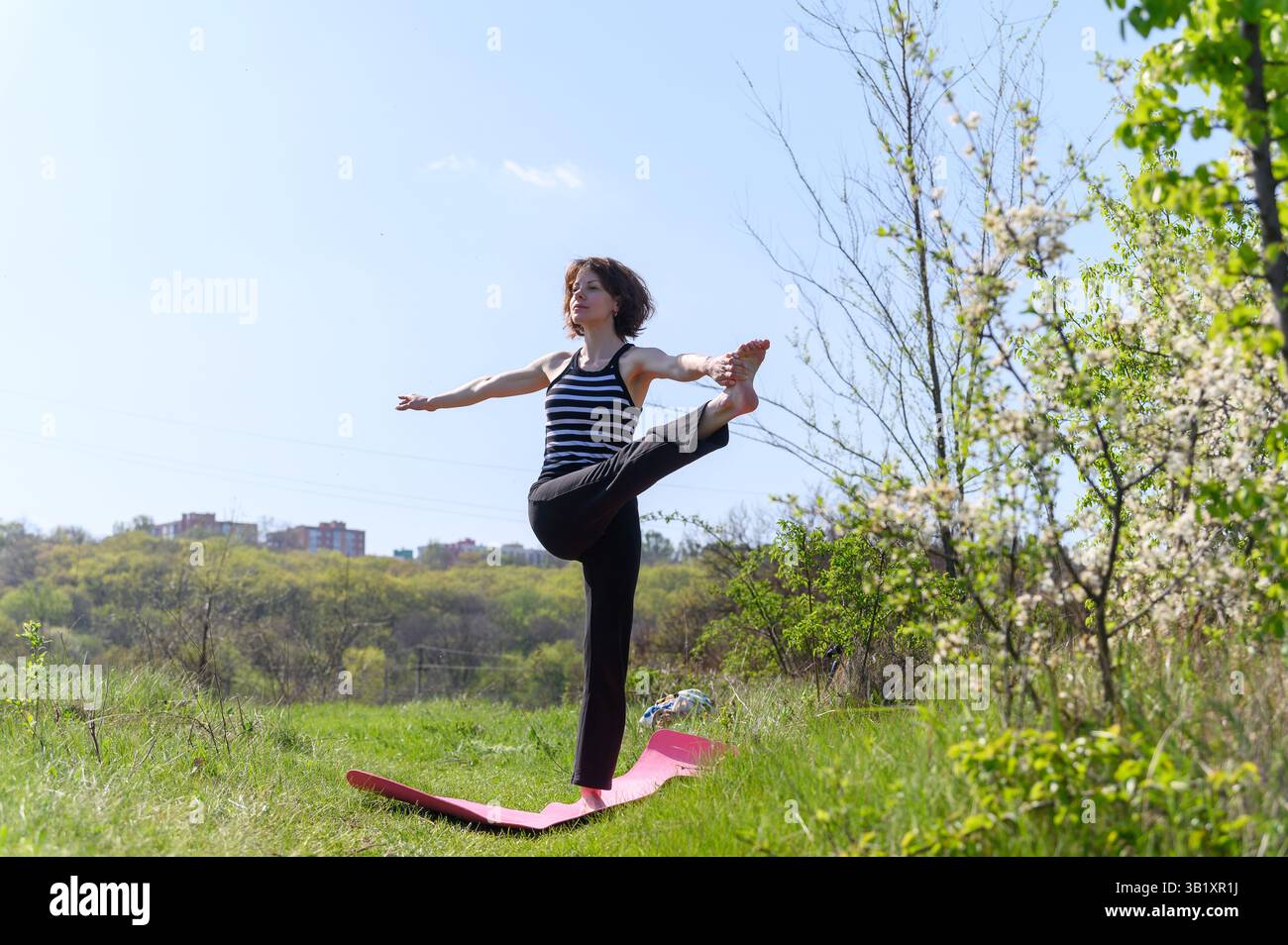 Frau balanciert in gestreckter Hand zu großer Zehenpose auf rosa Matte im grünen Feld, Arme gestreckt, Fernsicht, gestreifte Spitze, blühende Bäume, sonniger Tag Stockfoto