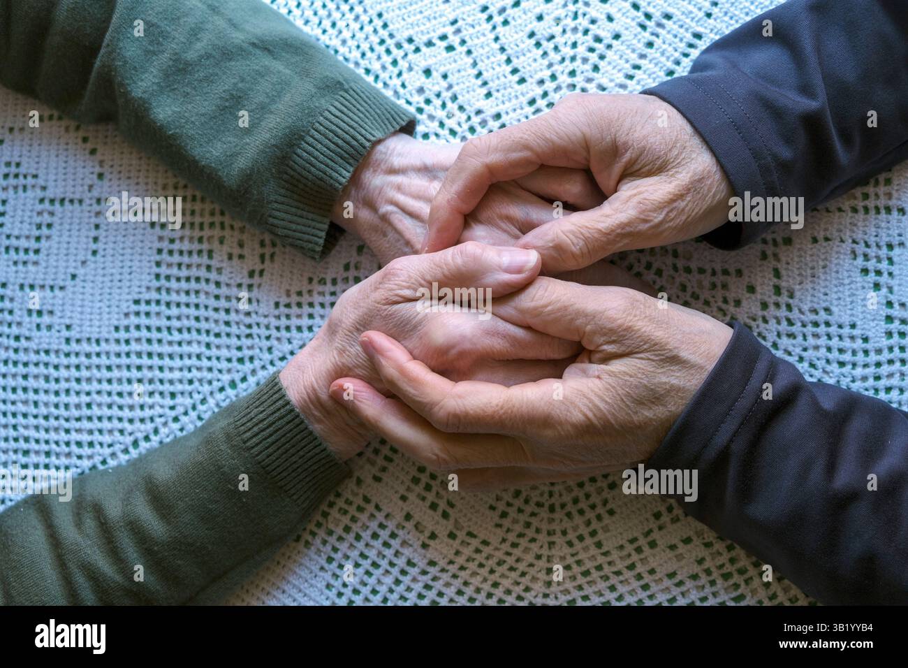 Zwei ältere Menschen, die die Hände fassen, symbolisieren Unterstützung, Fürsorge und emotionale Verbundenheit vor einem texturierten Stoffhintergrund. Stockfoto
