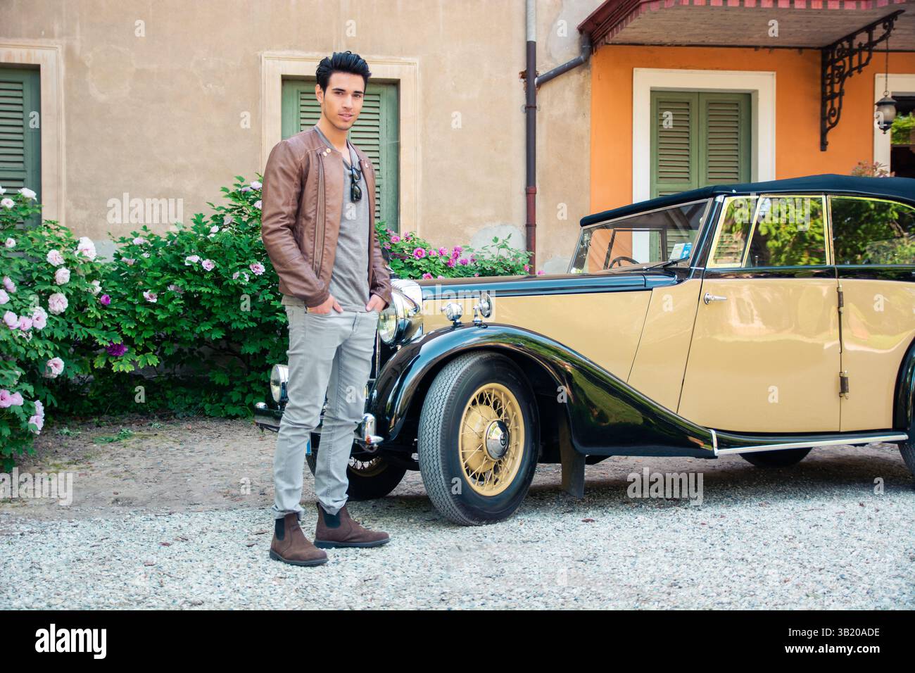 Ein junger Mann mit dunklen Haaren posiert selbstbewusst neben einem Oldtimer in einem bäuerlichen Garten voller Blumen. Die Umgebung bietet eine warme, sonnige Atmosphäre und charmante Architektur. Stockfoto