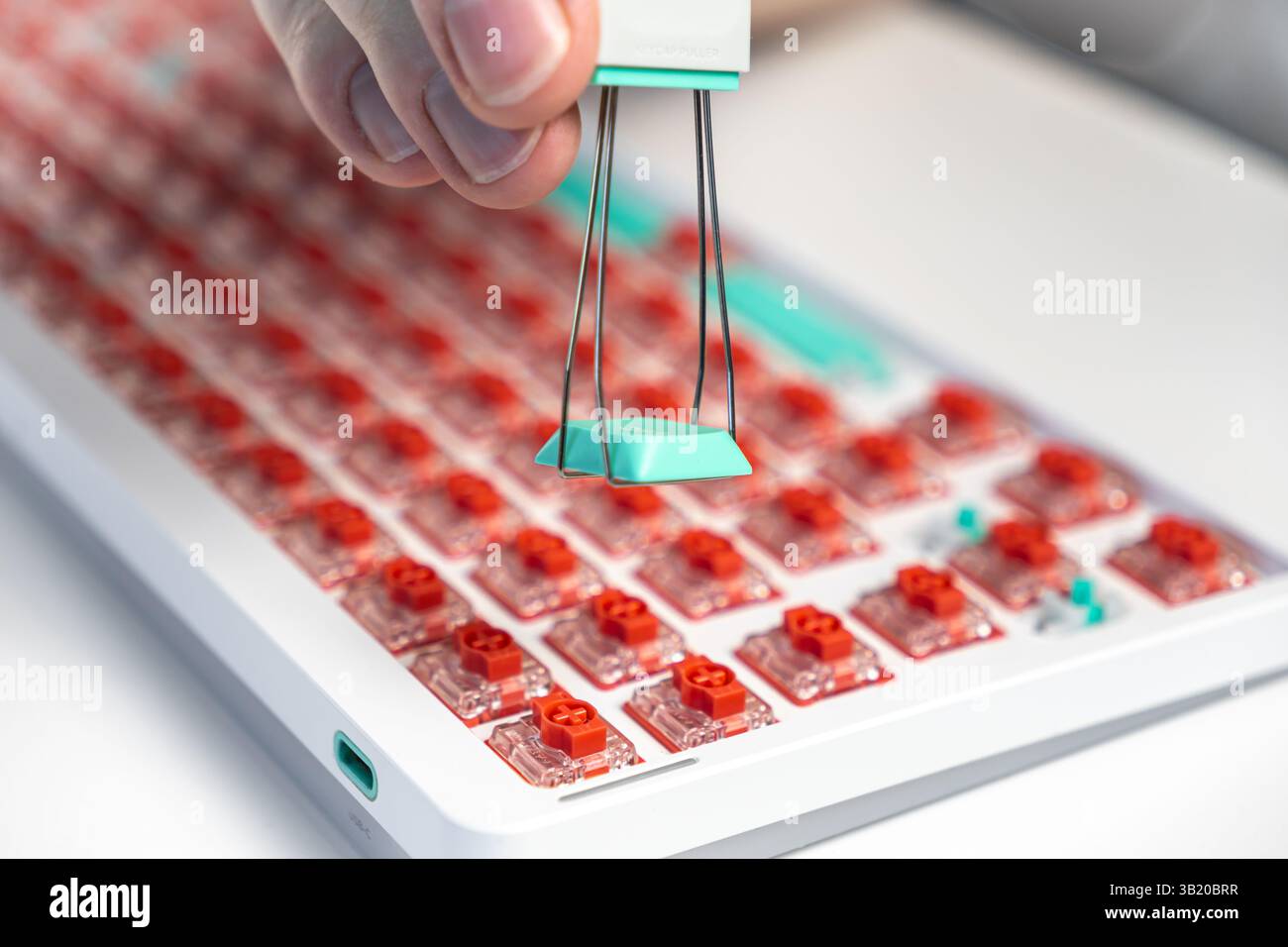 Makroansicht einer Hand, die mit einem Drahtzieher von einer kundenspezifischen mechanischen Tastatur mit roten Schaltern eine türkisfarbene Tastenkappe hebt. Hier wird die Reparatur von Heimwerkern und Technikern gezeigt Stockfoto