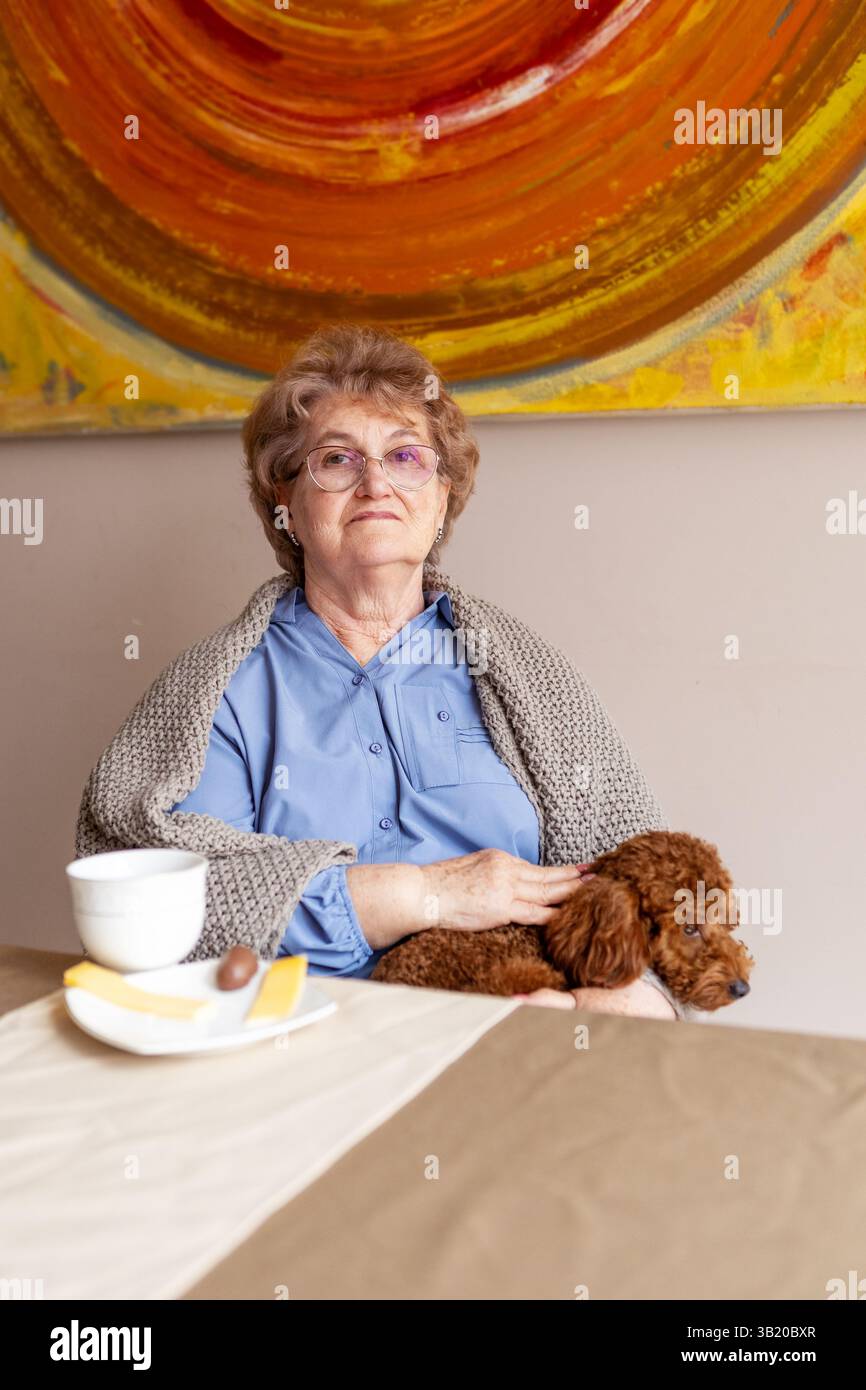 Einsame Seniorin sitzt zu Hause mit Hund, trinkt Tee und frühstückt, blickt nachdenklich in die Kamera, ruhiger Moment der Einsamkeit Stockfoto