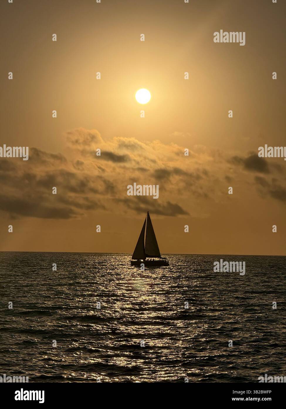Ein Segelboot gleitet über goldenes Wasser unter einem glühenden Sonnenuntergang und wirft eine ruhige Silhouette vor dem glitzernden Meer und dem traumhaften Himmel. - Smartphone-aufgenommenes Stockfoto