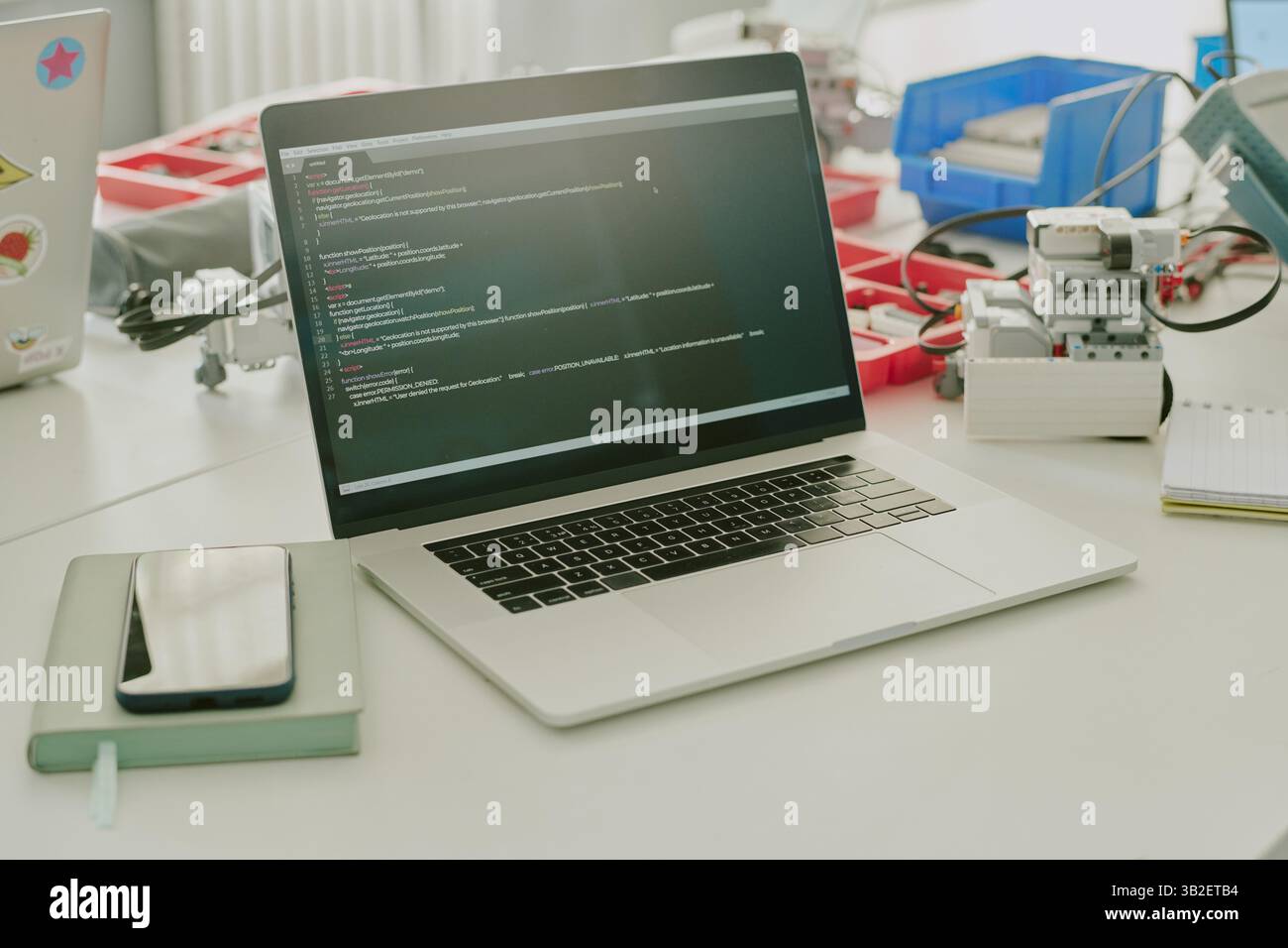 Mittelgroße Nahaufnahme eines silbernen Laptops mit Codes auf dem Bildschirm, Plastikkästen mit winzigen Details und elektronischem Gerät auf weißem Arbeitstisch Stockfoto