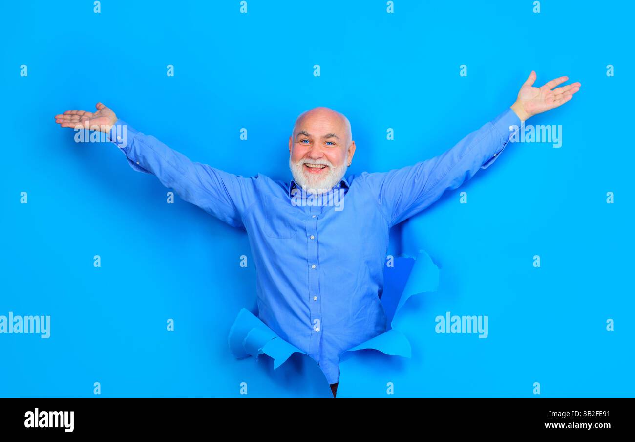 Lächelnder alter bärtiger Mann mit blauen Kontaktlinsen, der durch ein Loch im Papier schaut. Senior-Mann in stilvollem Hemd mit erhobenen Händen. Gutaussehender männlicher Blick Stockfoto