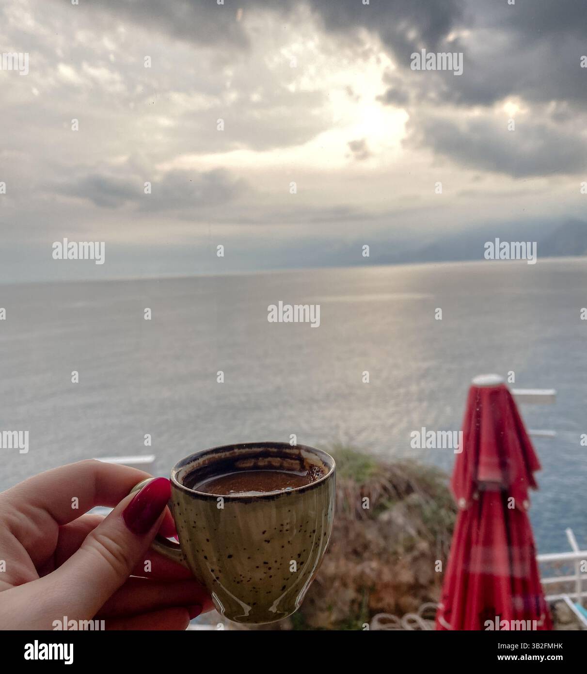 Eine Tasse Espresso wird an einem ruhigen Morgen unter bewölktem Himmel vor dem Meerblick gehalten. Stockfoto