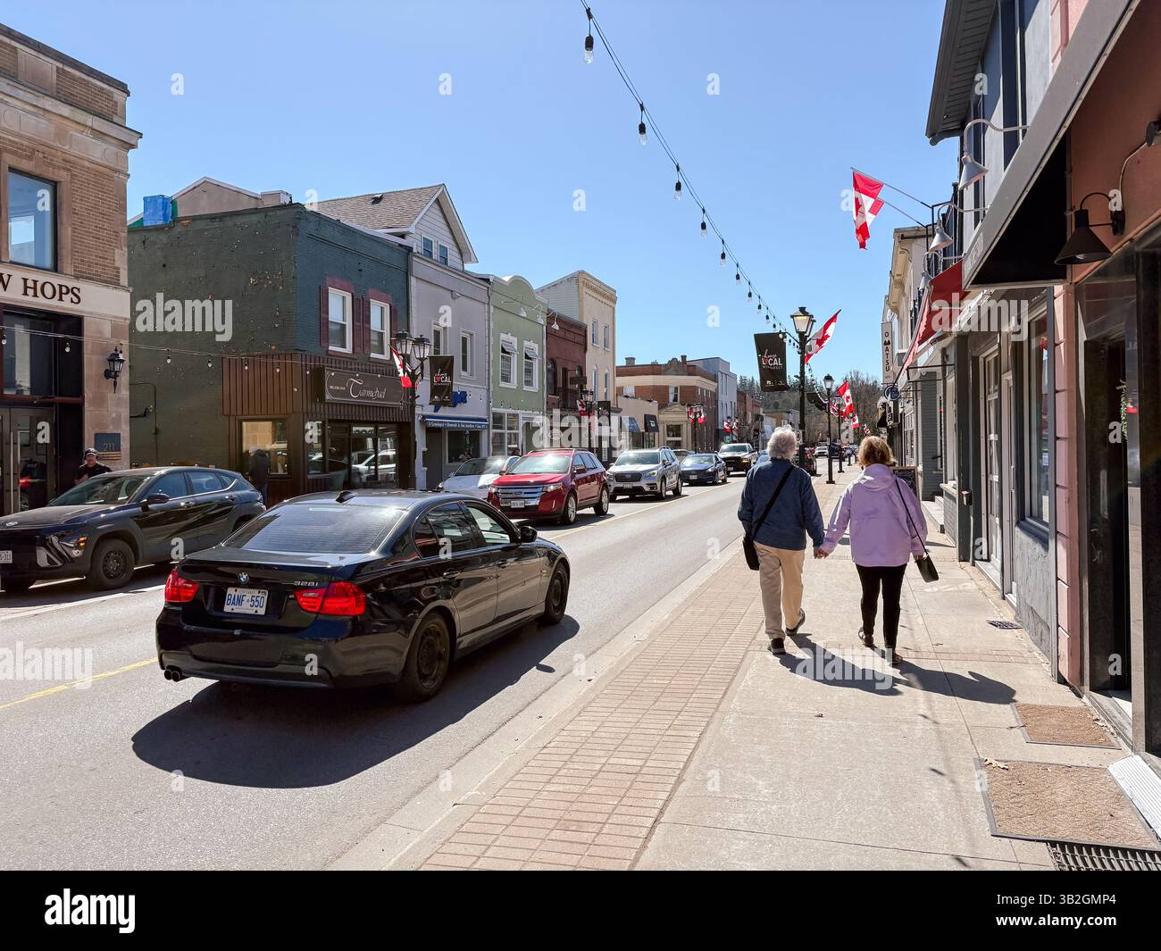 Ein sonniger Tag in Newmarket, Ontario, Kanada, zeigt die Hauptstraße mit Geschäften, Autos und Fußgängern. Auf der anderen Straßenseite hängen kanadische Fahnen. Stockfoto