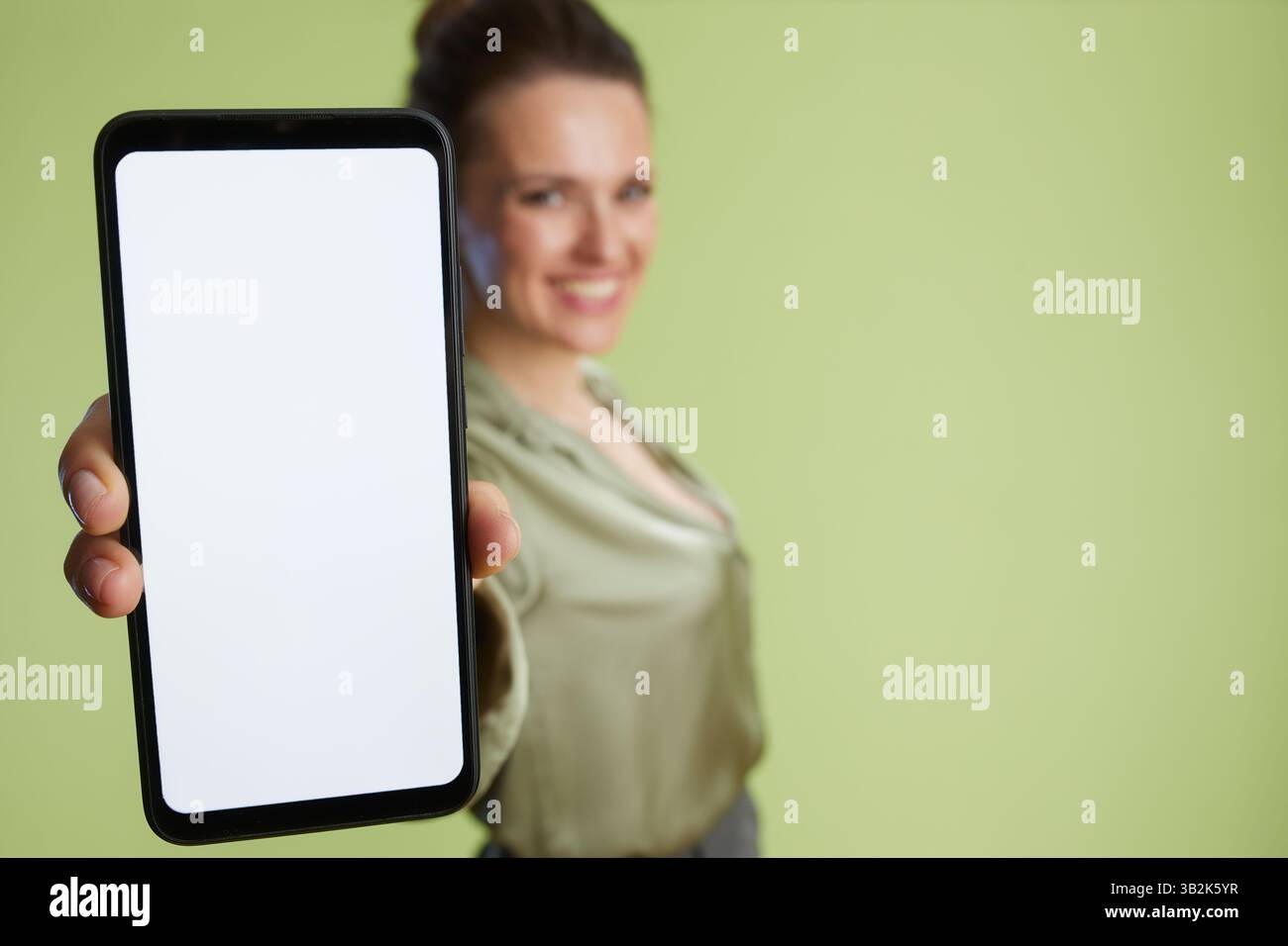 Eine lächelnde Frau hält ein schwarzes Smartphone mit einem leeren Bildschirm hoch. Förderung von Technologien mit weichem, hellgrünem Hintergrund. Stockfoto