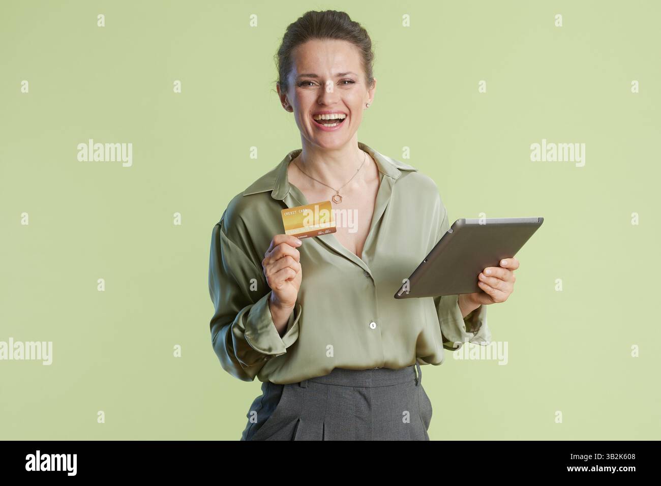 Die lächelnde Frau in den späten 30ern hält eine Kreditkarte und ein Tablet vor einem hellgrünen Hintergrund. Online-Shopping und die Einfachheit digitaler Produkte Stockfoto