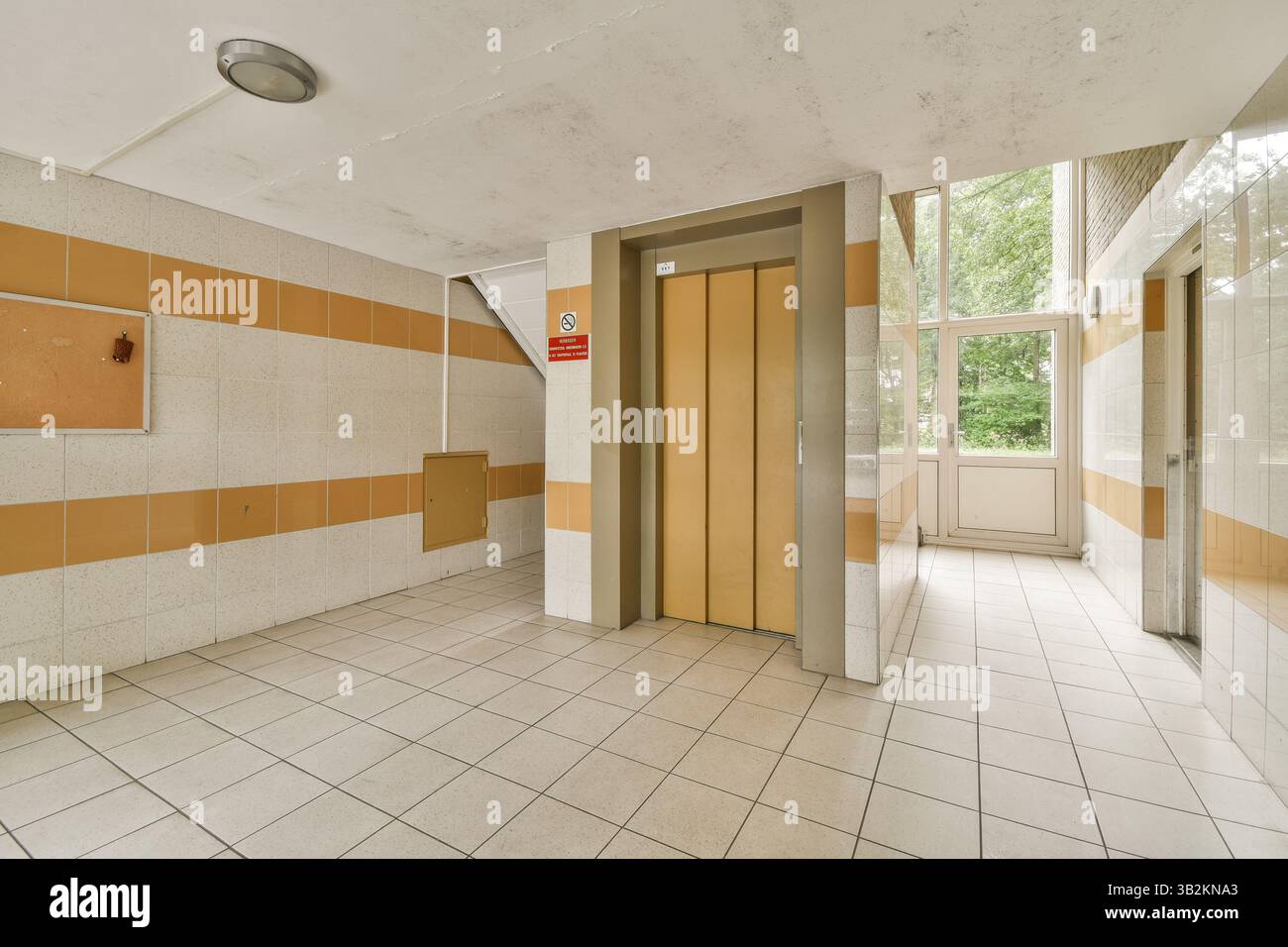 Blick auf den Innenraum mit einem modernen Flur mit gefliesten Wänden, neutralen Farben und Aufzug. Tageslicht beleuchtet den Raum durch große Fenster. Stockfoto