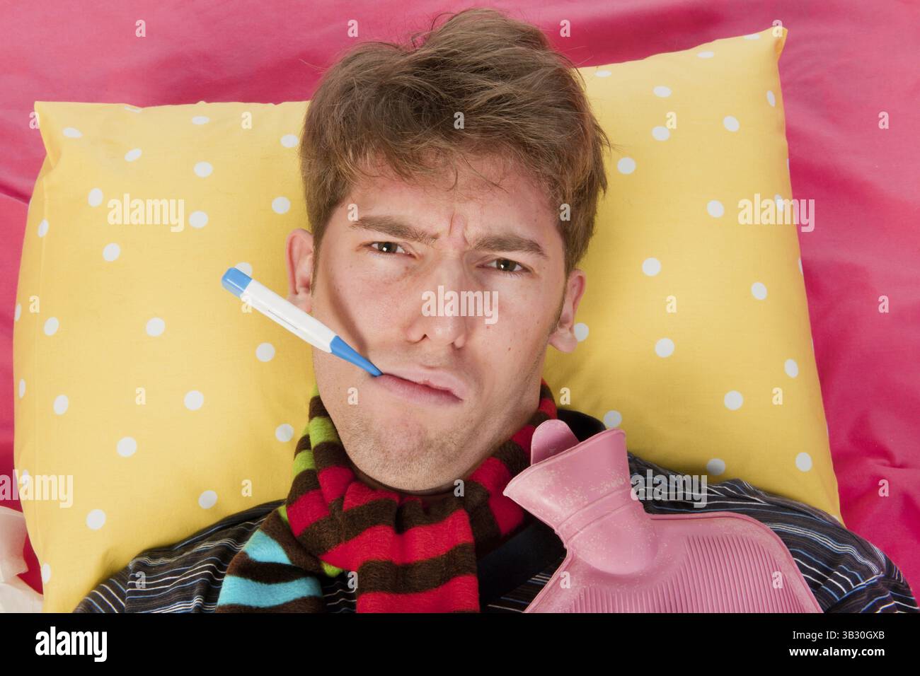 Ein sehr kranker Mann sitzt in seinem Bett mit einer Wärmflasche und einem Thermometer Stockfoto