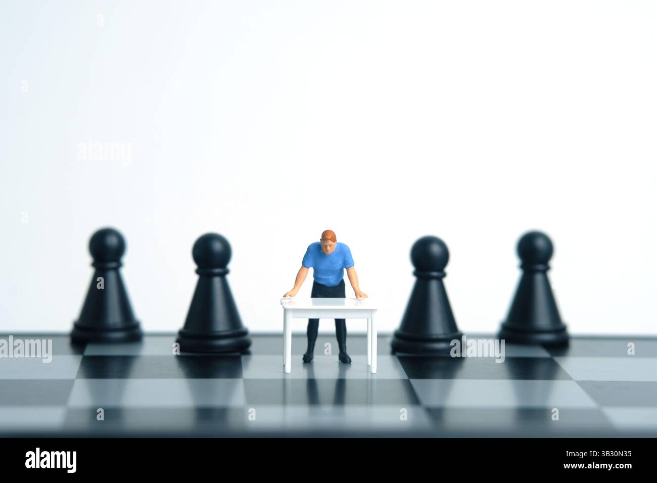 Ein Mann auf einem weißen Schreibtisch, der einen strategischen Plan über Schachbrett und Bauern denkt. Miniatur Tiny People Spielzeug Fotografie. Isoliert auf weißem Hintergrund. Bild Stockfoto