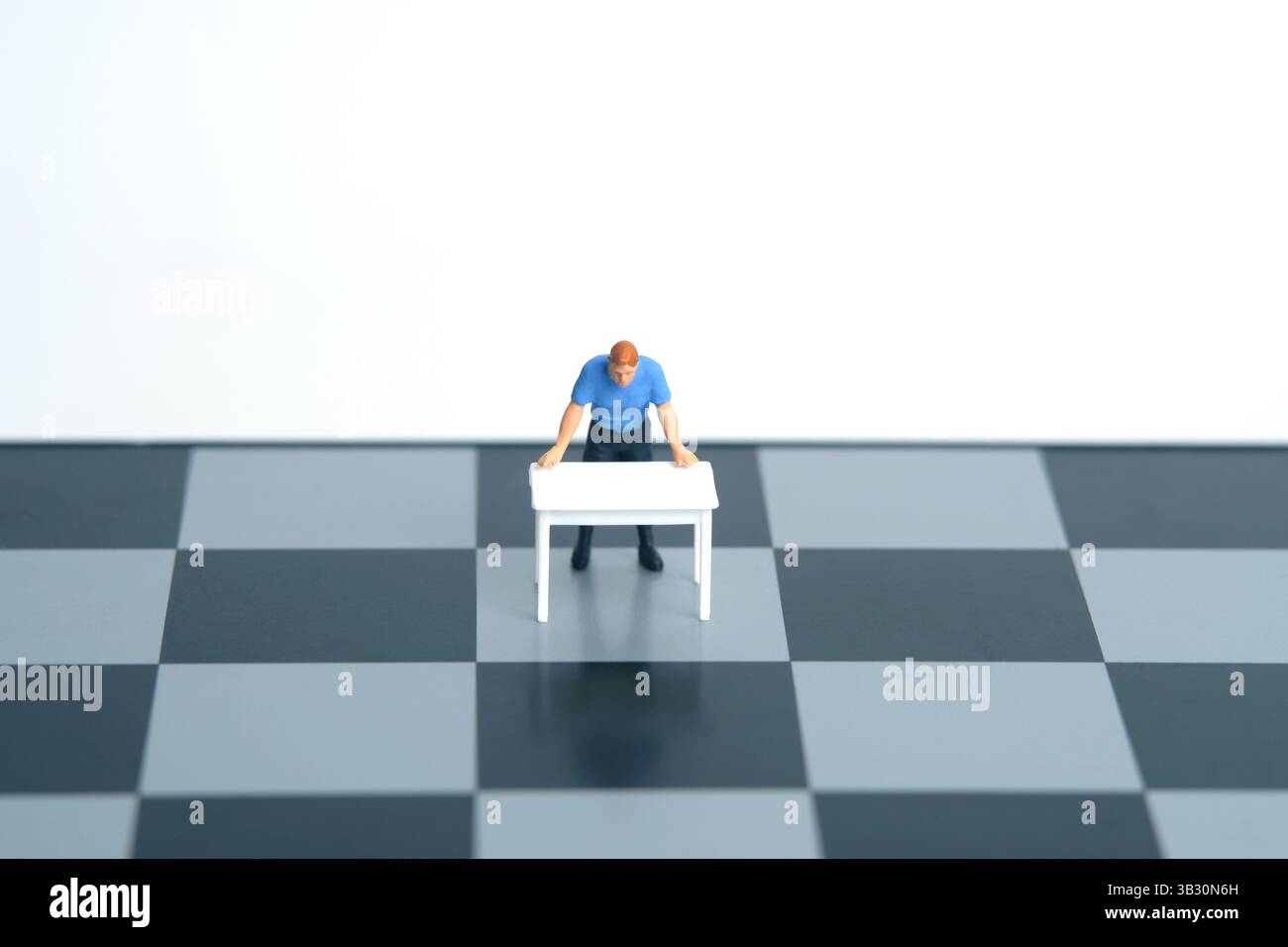 Ein Mann auf einem weißen Schreibtisch, der über dem Schachbrett einen strategischen Plan denkt. Miniatur Tiny People Spielzeug Fotografie. Isoliert auf weißem Hintergrund. Bild Foto Stockfoto