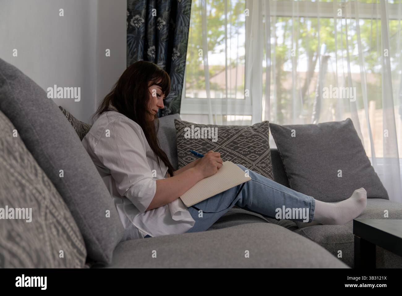 Die Schriftstellerin sitzt bequem auf dem Sofa in einem gemütlichen Wohnzimmer, schreibt Notizen für ihren neuen Roman und hält inspirierende Ideen fest Stockfoto