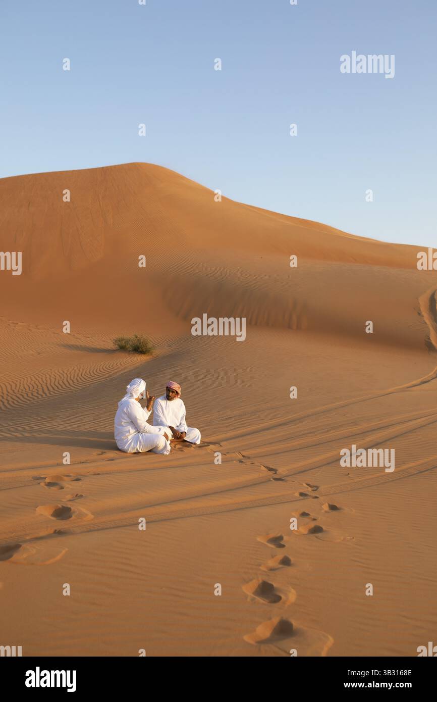Beduinenmänner in weißen Dschunddashas sitzen auf einer Sanddüne, Wahiba Sands Wüste oder Rimal Al Wahiba oder Sharqiyya Sands, Janub Ash-Sharqiyya Region, Oman, A Stockfoto