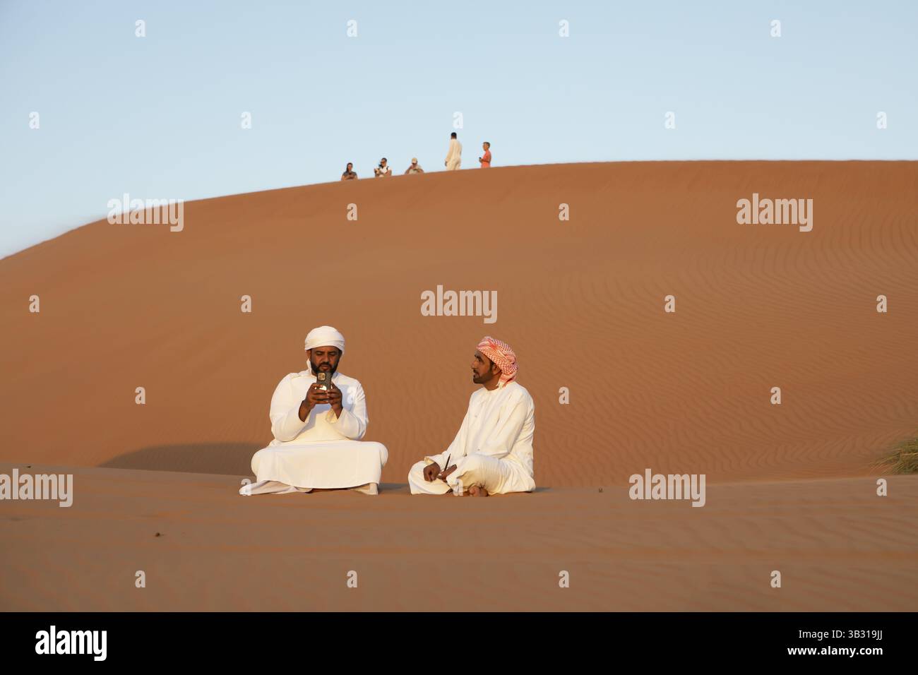 Beduinenmänner in weißen Dschunddashas sitzen auf einer Sanddüne, Wahiba Sands Wüste oder Rimal Al Wahiba oder Sharqiyya Sands, Janub Ash-Sharqiyya Region, Oman, A Stockfoto